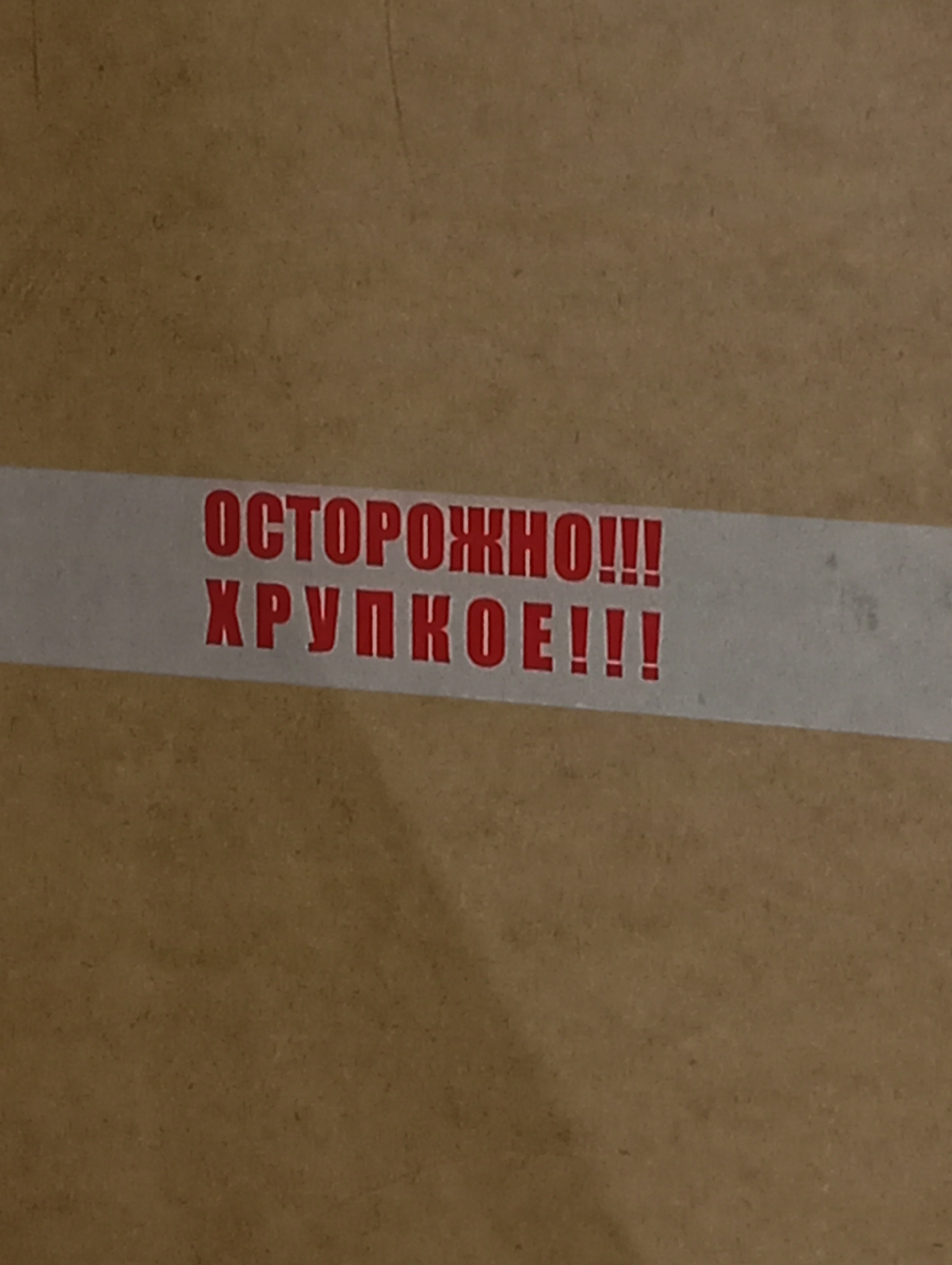 A close-up shot captures a section of a cardboard box, marked with a white packing tape. Printed on the tape in red, Cyrillic letters are the words "ОСТОРОЖНО!!!" and "ХРУПКОЕ!!!", which translate to "CAUTION!!!" and "FRAGILE!!!" respectively. The tape appears slightly aged, with some discoloration and minor blemishes. The cardboard is a medium brown color with a visible texture. No location or city is discernible from the image.