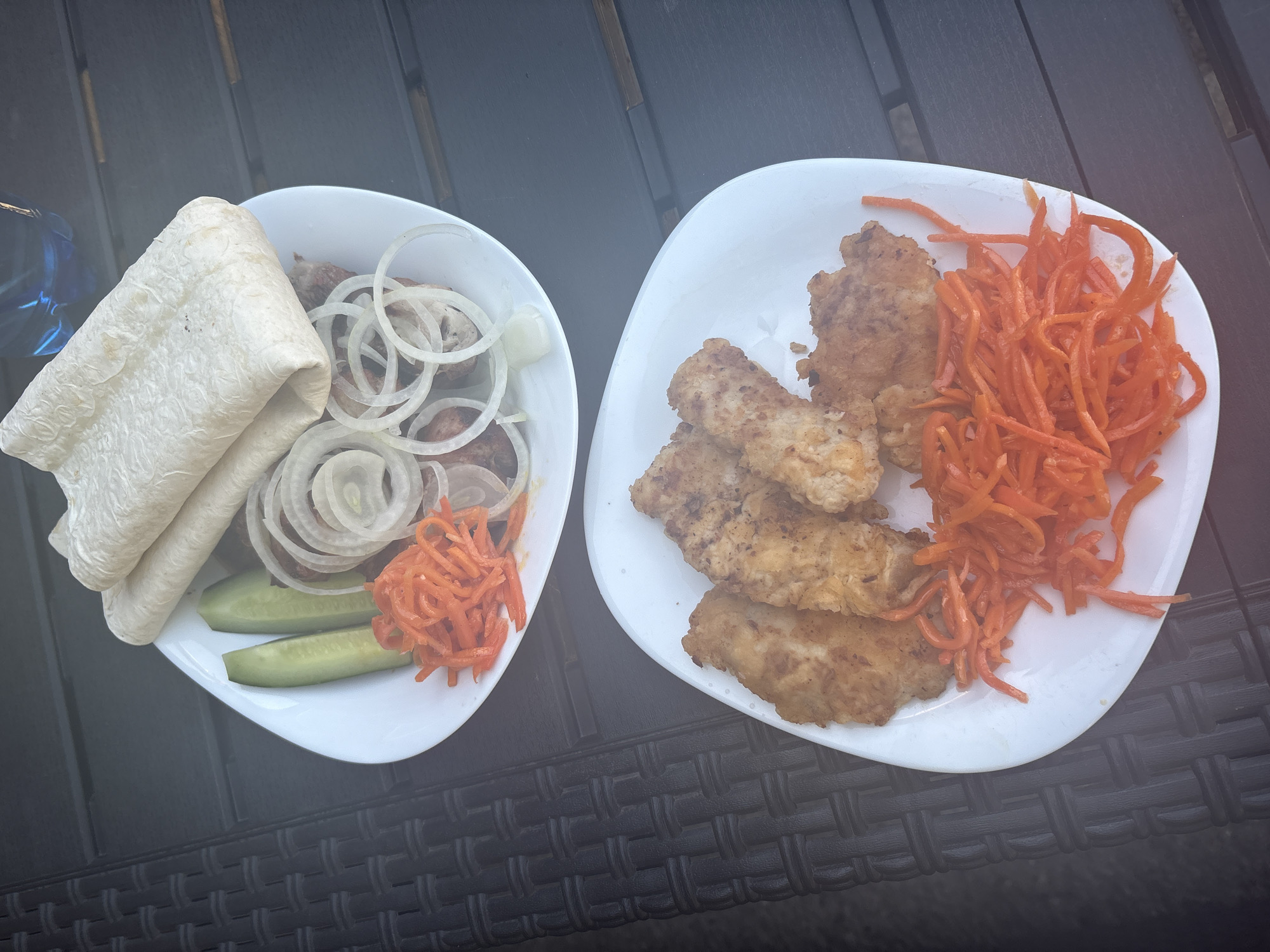 Two white plates of food are presented on a dark, textured surface that appears to be an outdoor table.
The top plate contains a serving of thinly sliced meat, possibly grilled, accompanied by several rings of raw white onion, two cucumber spears, shredded orange carrots, and two folded pieces of flatbread.
The bottom plate features several pieces of breaded and fried chicken tenders, served with a generous portion of shredded orange carrots. The food is arranged on the plates in a visually appealing manner, suggesting it is ready to be eaten. The background is slightly out of focus, but a blue object, possibly a bottle of water, is visible in the upper right corner. The scene is set in the city of Kommunarka, Russia.