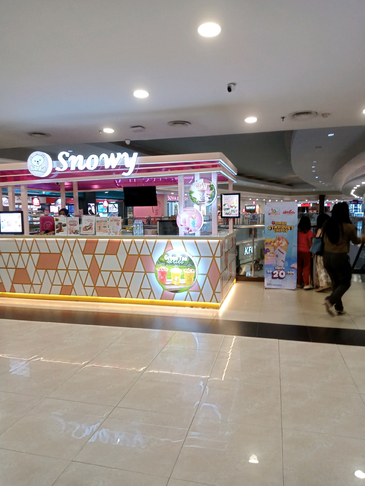 The image shows a "Snowy" branded dessert and drink kiosk in a shopping mall. The kiosk has a pink and white color scheme and features a logo with a teddy bear. Display screens show images of the products, including milkshakes and ice cream. A banner advertising "Dimsum Simba Stardust Crepes" is visible to the right of the kiosk, featuring a price of "RP 20.000". Several people are present in the scene, including a person behind the counter at the kiosk, and three people standing near the banner. The flooring is tiled and reflective. The image was taken in Cirebon, Indonesia, likely at a mall such as Grage City Mall or CSB Mall, as these are common locations for such kiosks and brands.