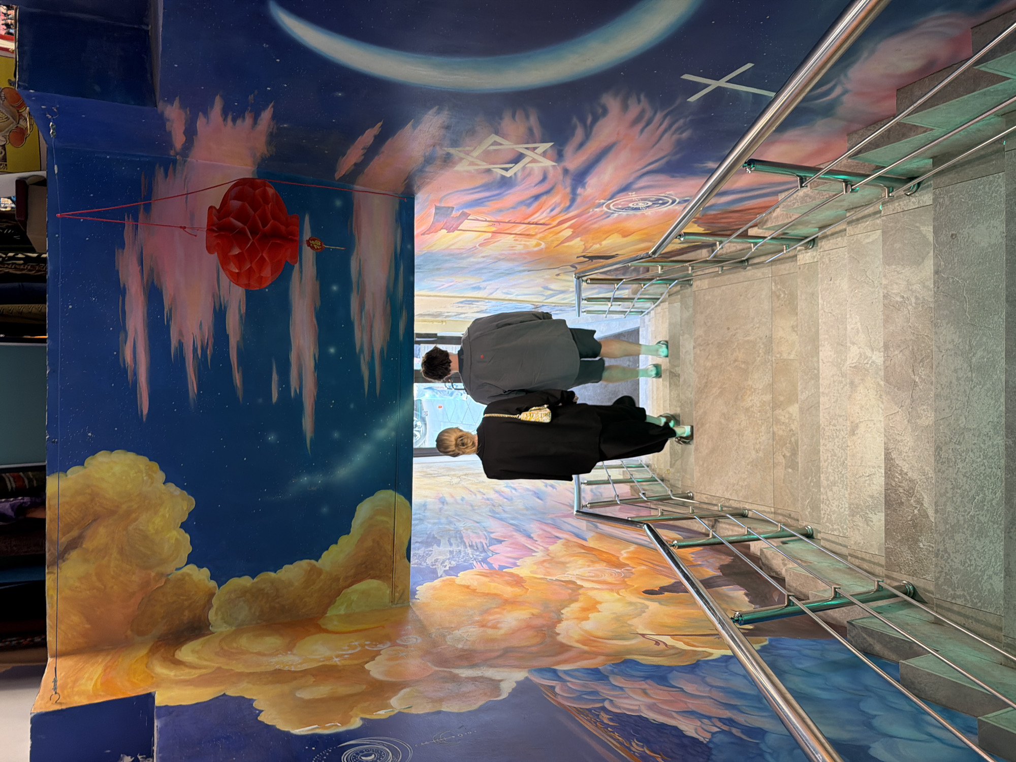 Here is a description of the image:
Location: Moscow, Russia, in the Museum of Jewish History and Tolerance.
Environment: The image captures an interior space of the museum featuring a hallway or pathway with murals painted on the walls and ceiling. The artwork depicts a celestial or spiritual theme with clouds, stars, and cosmological symbols. A staircase with metal railings leads towards the painted artwork.
People: There are two people present in the image. Both of them appear to be walking along the path with their backs to the camera.
Objects: The primary objects in the image include the painted murals, metal railings of the staircase, steps of the staircase, and a hanging red decorative element.
Actions: The two individuals are walking through the museum space.
Interactions: The interaction between the individuals and the environment is that they are moving through it, presumably observing or exploring the murals and architectural elements.
