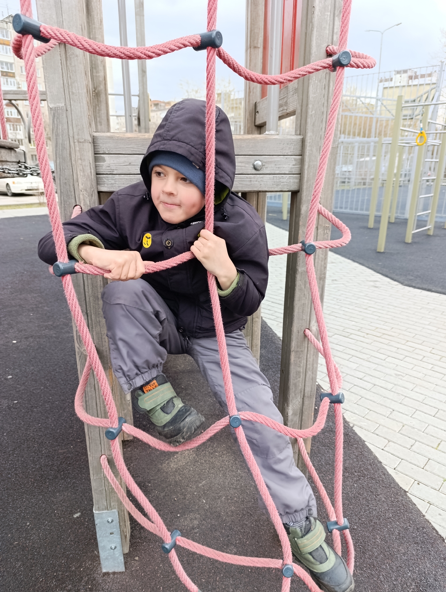 A young boy wearing a black hooded jacket, gray pants, and dark-colored boots with green accents is on a playground in Perm, Russia. He is climbing on a wooden structure with a pink rope net. The structure appears to be part of a climbing frame or jungle gym. The boy is holding onto the ropes with both hands. In the background, there are other playground structures, buildings, and a cloudy sky. The ground is covered in paving stones and black rubber padding.
