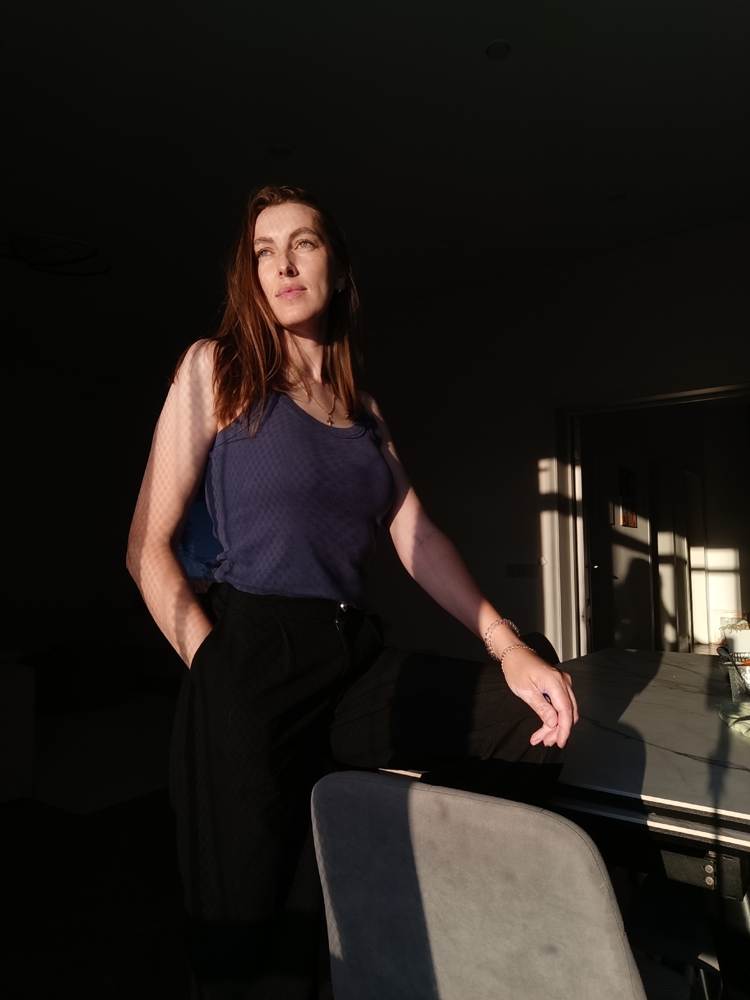 A woman with long, brown hair, wearing a blue tank top and black pants, stands in a dimly lit room, illuminated by a streak of sunlight. Her left hand is in her pocket, and her right arm rests on a table, adorned with a bracelet. The sunlight casts a patterned shadow on her arm and chest, and also falls across a doorway and wall in the background. The room appears to be an indoor space, possibly an apartment or office, with a table and chairs visible. The lighting suggests it is either early morning or late afternoon. There is no specific indication of Chaikovsky, Russia in the image itself.