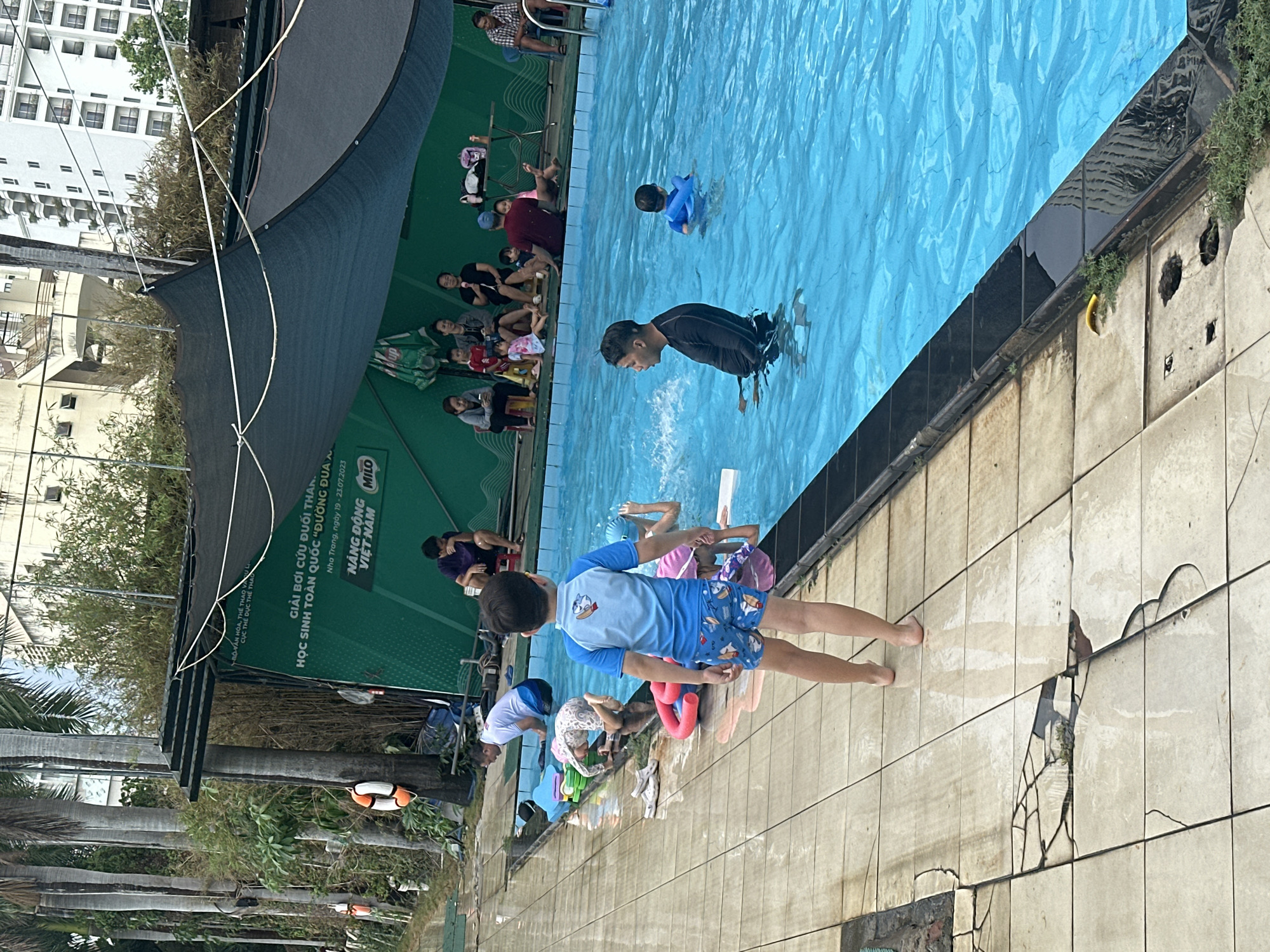 An outdoor swimming pool scene in Nha Trang, Vietnam, depicts a vibrant swimming event or program for students. The image, captured during daytime under a clear sky, shows numerous children and adults engaged in activities in and around a large, bright blue swimming pool.
In the pool, a man in a dark rashguard stands in the shallow end, while a young child in a blue swim shirt splashes in the water. Several other children are visible swimming or standing in the pool. On the tiled deck surrounding the pool, a boy in a blue rashguard and patterned shorts stands near the edge, observing. Other children are seen playing or preparing to enter the water, some with swimming aids. A group of adults and children are seated on benches along a green wall on one side of the pool, seemingly watching the activities.
The setting includes a dark mesh shade canopy overhead. In the background, a multi-story building and lush green vegetation, including palm trees, are visible. The pool deck is made of light-colored tiles, some of which are visibly cracked or damaged.
A prominent green banner along the poolside displays Vietnamese text, detailing the event: "GIẢI BƠI CỨU ĐUỐI THANH THIẾU NIÊN HỌC SINH TOÀN QUỐC “ĐƯỜNG ĐUA XANH”" (Youth Drowning Prevention Swimming Competition, National Student "Green Race"), held in "Nha Trang, ngày 19 - 23.07.2023". The banner also mentions "NĂNG ĐỘNG MILO" (Dynamic Milo) and is associated with the "BỘ VĂN HÓA, THỂ THAO VÀ DU LỊCH" (Ministry of Culture, Sports and Tourism) and "CỤC THỂ DỤC THỂ THAO" (Sports and Physical Training Department), indicating an officially organized event.