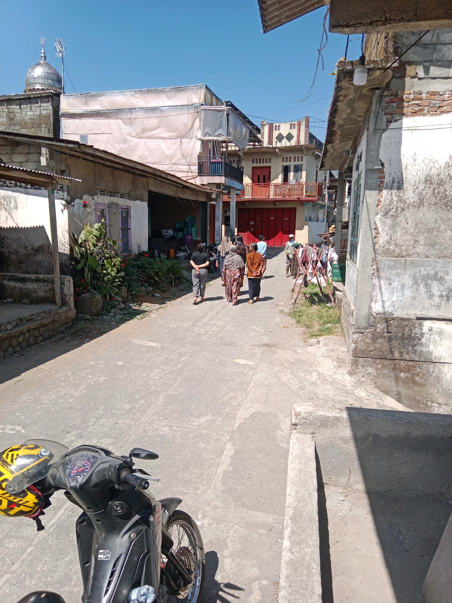 In Jurangjeru, Indonesia, a sunny outdoor scene captures a narrow, paved street lined with buildings. On the left, a mosque with a silver dome and antenna is visible, adjacent to a weathered, light-colored structure with a corrugated metal roof. Further down the street, a building with a light pink facade and balconies stands between two other structures, one with red doors. On the right side of the street, a rough, gray building with visible brickwork and exposed concrete dominates the foreground. A small patch of grass with laundry hanging on a rack is visible further down. Several individuals, some in traditional Indonesian attire, are walking in groups along the street. In the foreground, a black motorcycle with a yellow and black helmet is parked. The overall impression is one of a lived-in, communal urban environment on a clear day.