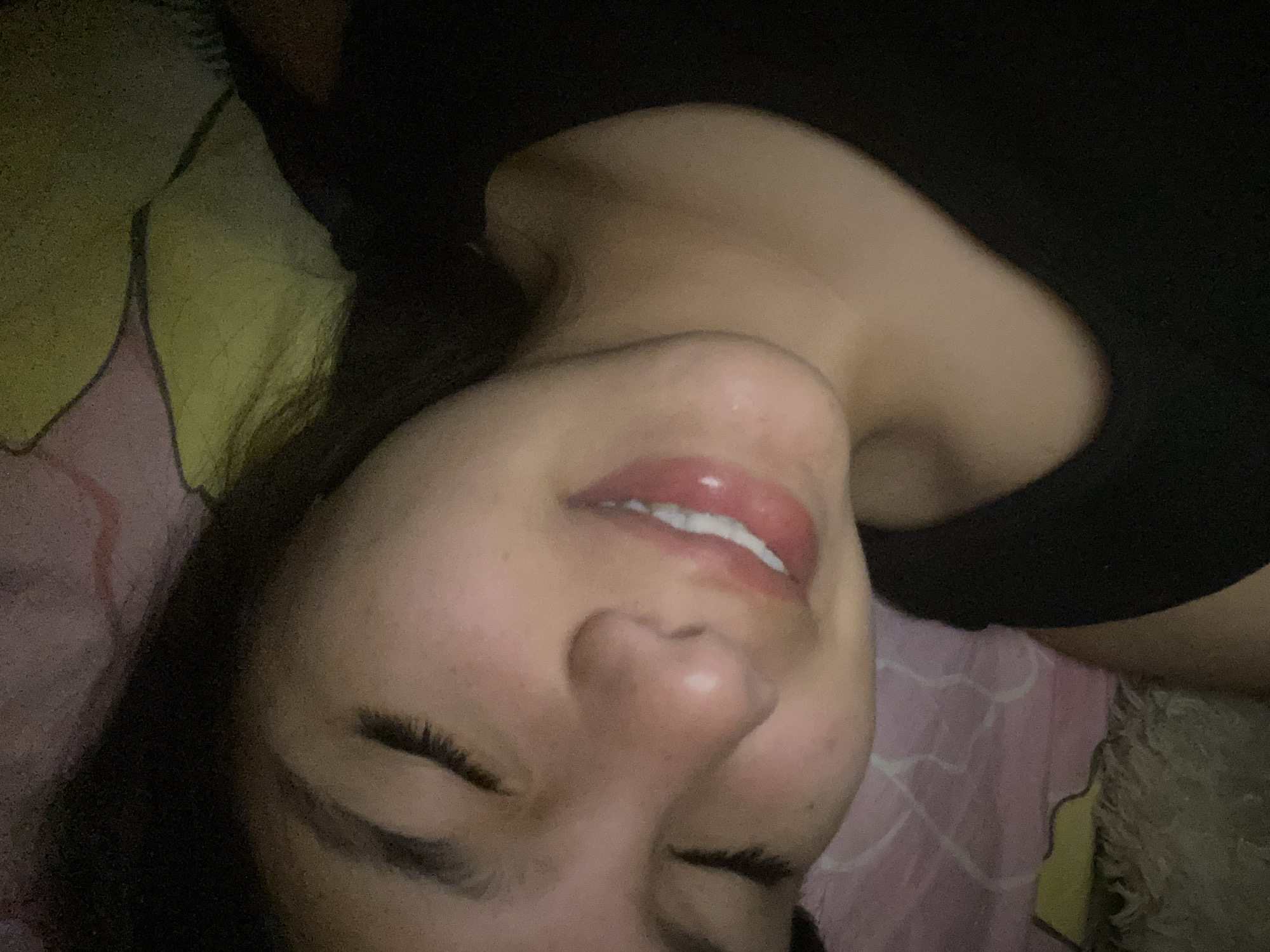 A close-up, underexposed image shows a person lying on patterned bedding, possibly in an interior setting. The individual has dark hair and closed eyes, with lips slightly parted revealing teeth. The person is wearing a dark-colored top. The background includes a yellow and pink patterned sheet. No information indicates the location is in Cotoperíz, Venezuela.
