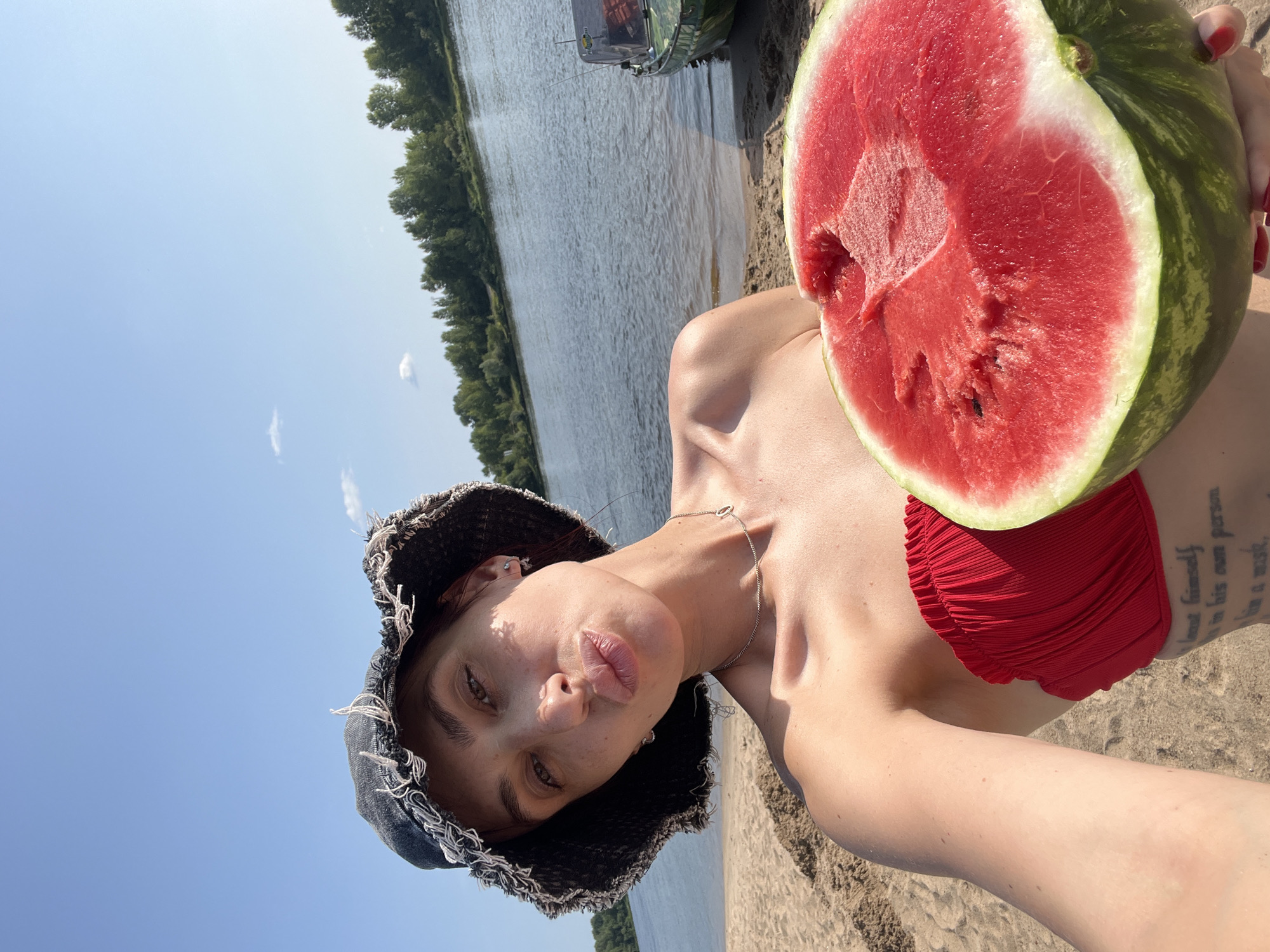 A woman with freckles on her face and shoulders, wearing a denim bucket hat and a red strapless bikini top, holds a half-eaten watermelon while posing for a selfie. She is on a sandy beach with a calm river and a tree line in the background. A small boat is visible docked on the right side of the river. The sky is clear blue with a few scattered clouds. The woman has tattoos on her upper torso. The scene appears to be in the city of Nizhny Novgorod, Russia, during a warm, sunny day.