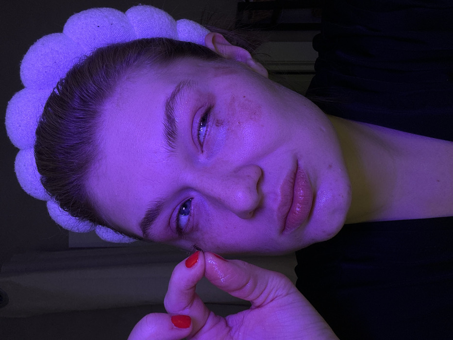 A woman with fair skin and blue eyes tilts her head in a close-up shot with purple lighting. She's wearing a white headband. Her expression is serene. She holds a small object between her finger and thumb, coated in liquid. Red nail polish adorns her nails. A dark-colored garment is visible near her neck. Some freckles or blemishes are visible near her eye.