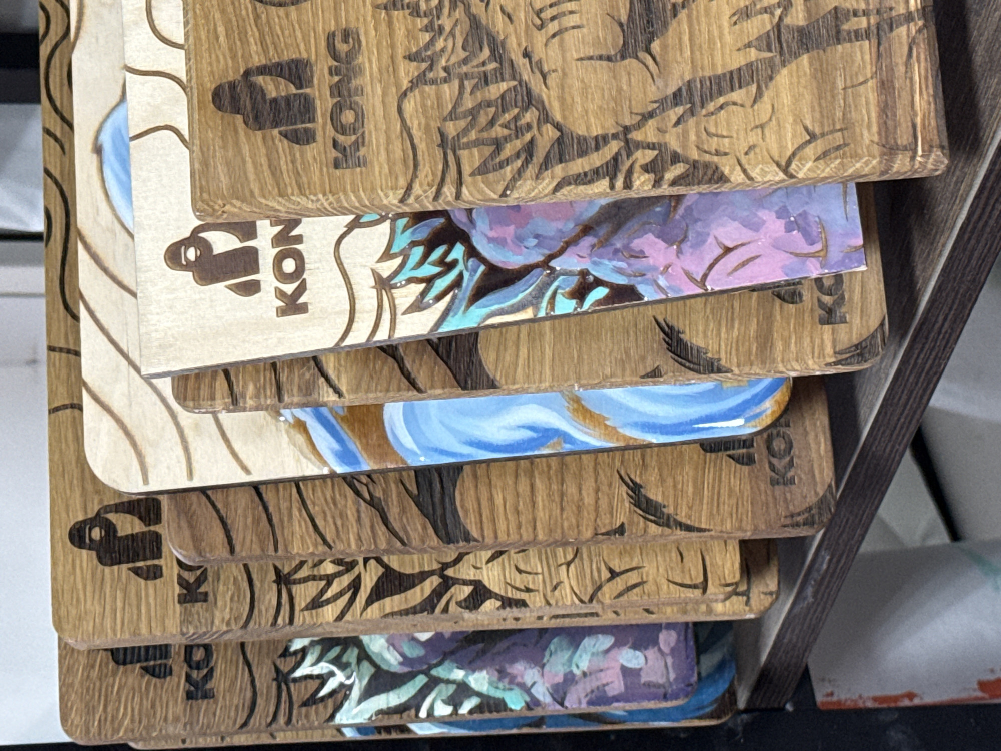 A stack of wooden items, possibly coasters or small plaques, rests inside a dark-colored wooden tray. The items are printed with the word "KONG" and images of what appear to be cartoon apes and leaves. The image contains the coordinates of Chaikovsky, Russia. No people are visible in the scene.