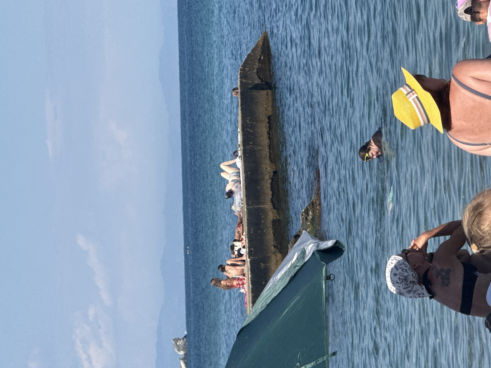 The image depicts a sunny day at a coastal area, likely a beach or harbor in Partenit, Ukraine. In the foreground, partially obscured by a green umbrella, are two individuals facing away from the camera. The person on the left is wearing a headscarf and sunglasses, with a tattoo visible on their back. The person on the right is wearing a yellow straw hat.
In the mid-ground, a concrete pier extends into the turquoise sea. Several people are sunbathing and relaxing on the pier. Two individuals are seated with their backs to the camera, and another is lying down. Further out in the water, a person wearing swimming goggles is visible.
In the background, the blue sea stretches to the horizon, meeting a hazy blue sky with scattered clouds. Distant, undulating hills form the coastline. A rocky outcrop is visible on the left side of the image, near the distant shore. The overall impression is one of a relaxed summer day by the sea.