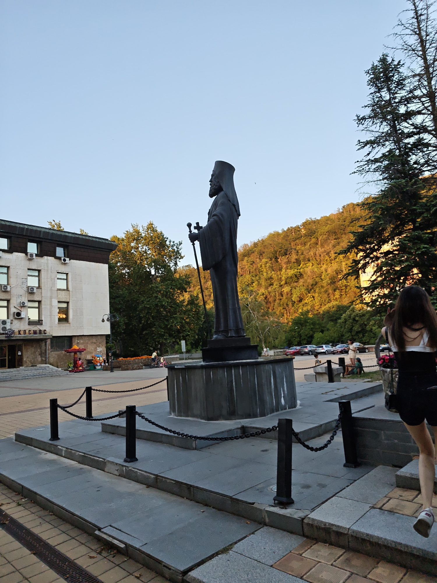 The image shows a statue of a man in religious attire, holding a staff, in an outdoor plaza. The statue is located in front of a building with the word "КОСМОС" visible on its facade, which is the "Kosmos" restaurant and hotel complex in Lovech, Bulgaria. A young woman with long dark hair is walking up the steps to the statue. In the background, there is a tree-covered hill bathed in the warm light of what appears to be sunset. The plaza is paved with stone and has black chain barriers around the statue's base. Other people are visible in the distance, including children on a small amusement ride.