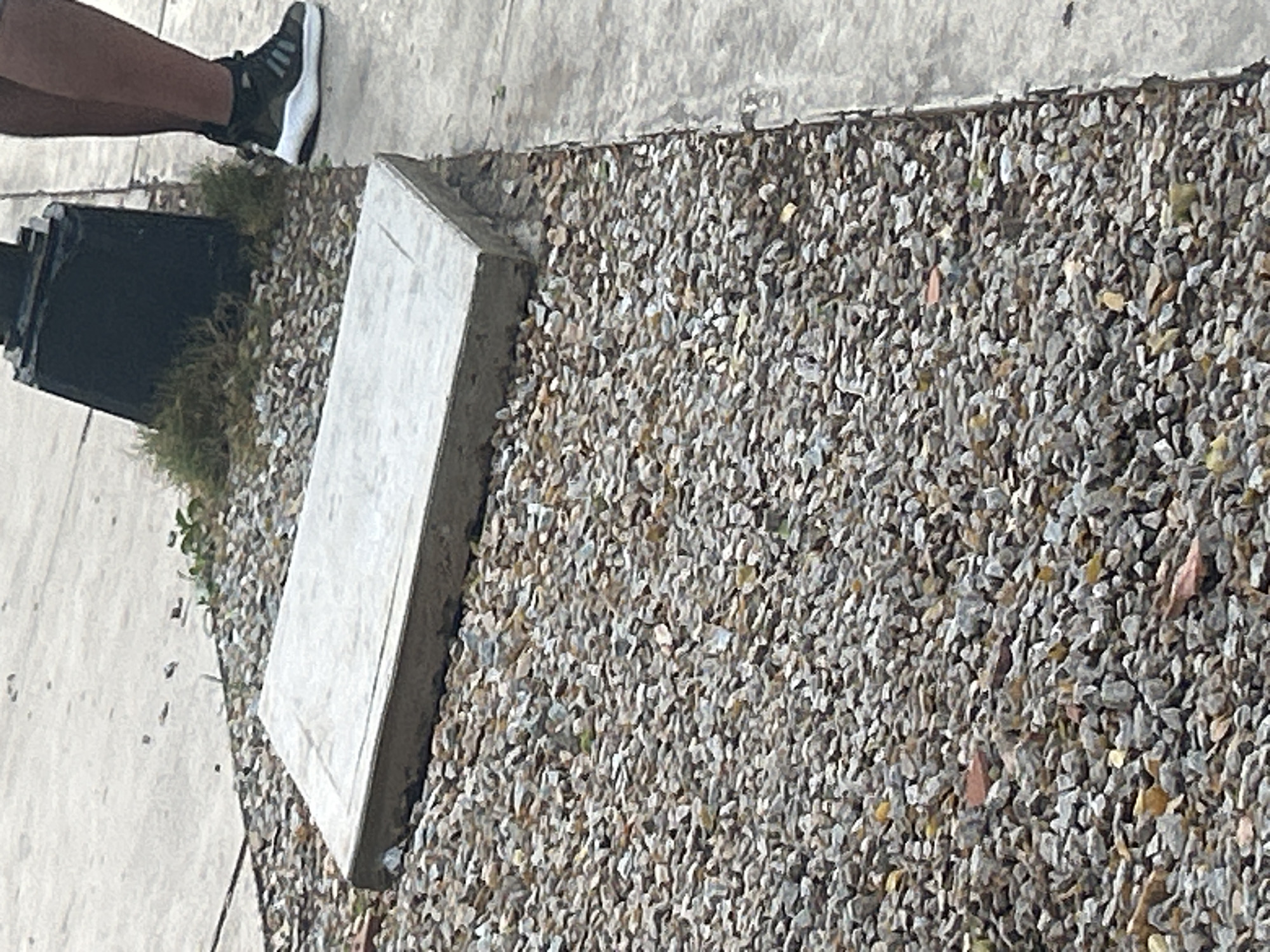 The image shows a concrete slab lying on a bed of small, gray and brown gravel, adjacent to a concrete sidewalk. To the right of the sidewalk, a person's legs and feet are visible, wearing dark athletic shoes with white soles. Behind the gravel and slab, a black metal pole, possibly part of a lamppost, stands amidst sparse green vegetation. The scene appears to be an outdoor urban environment, likely in Santa Ana, Venezuela.