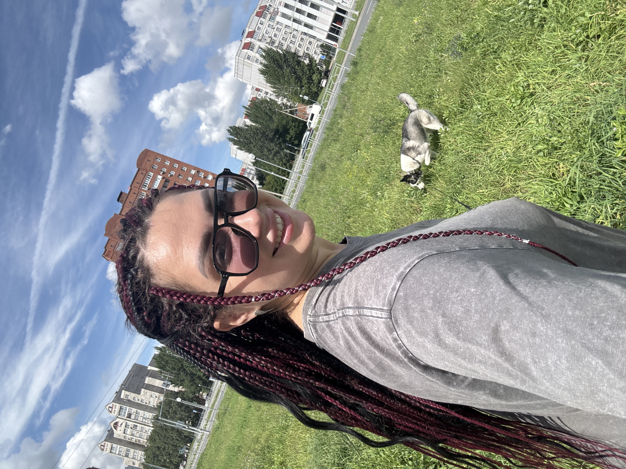 A woman with dark hair in braids, wearing sunglasses and a gray t-shirt, smiles at the camera in an outdoor setting. Behind her, a husky-like dog is running in a grassy field. In the background, there are apartment buildings and trees, suggesting an urban environment. The sky is blue with white clouds. The scene appears to be from the city of Tolyatti, Russia, potentially in a park or green space near residential areas.