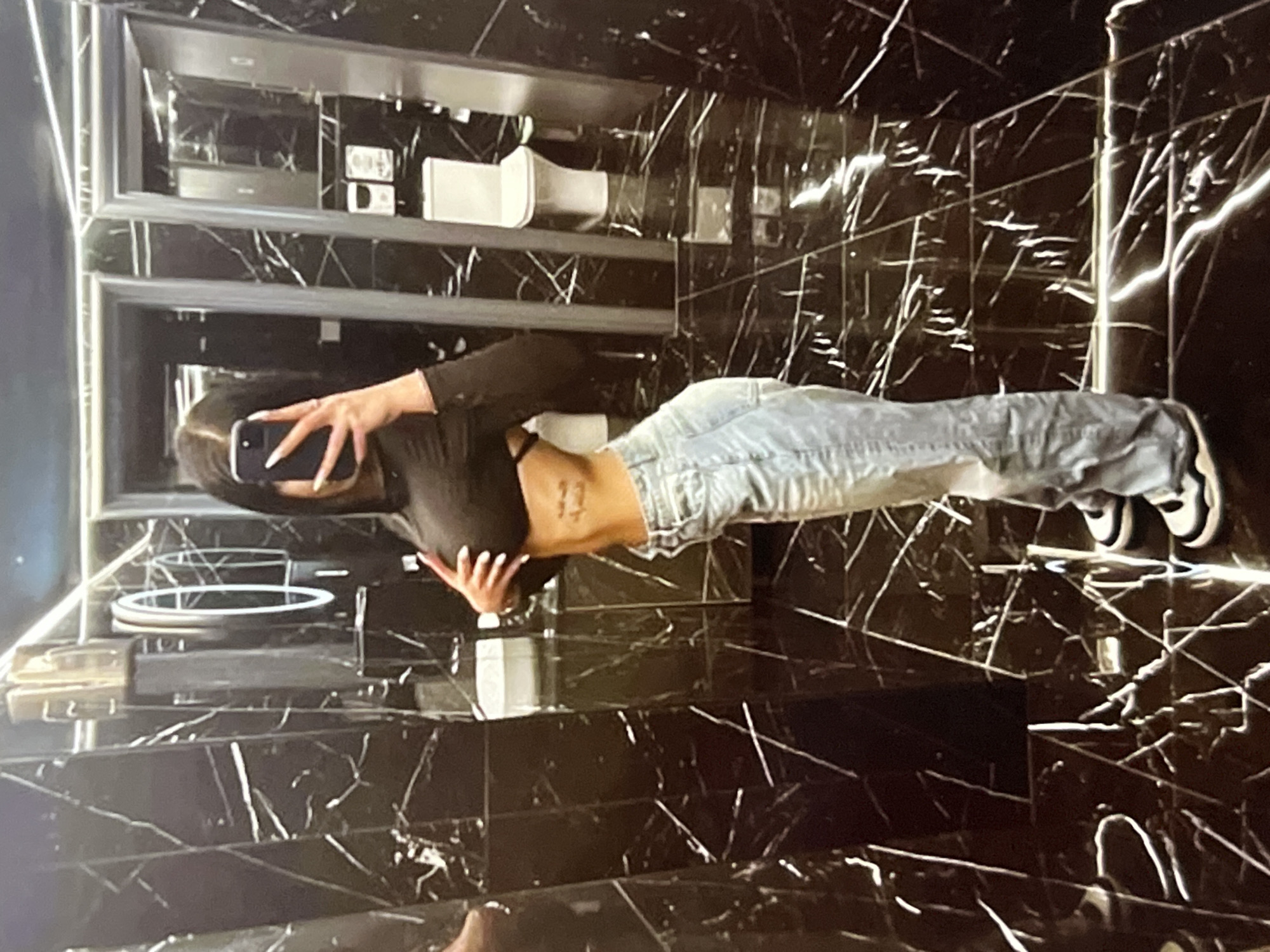 A woman is taking a mirror selfie in a modern, black marble-tiled bathroom. She is wearing a black crop top and light-wash baggy jeans. A tattoo is visible on her side. The bathroom features multiple mirrors, stalls, a white toilet, and a sink. The lighting is dim, creating reflections on the glossy surfaces.