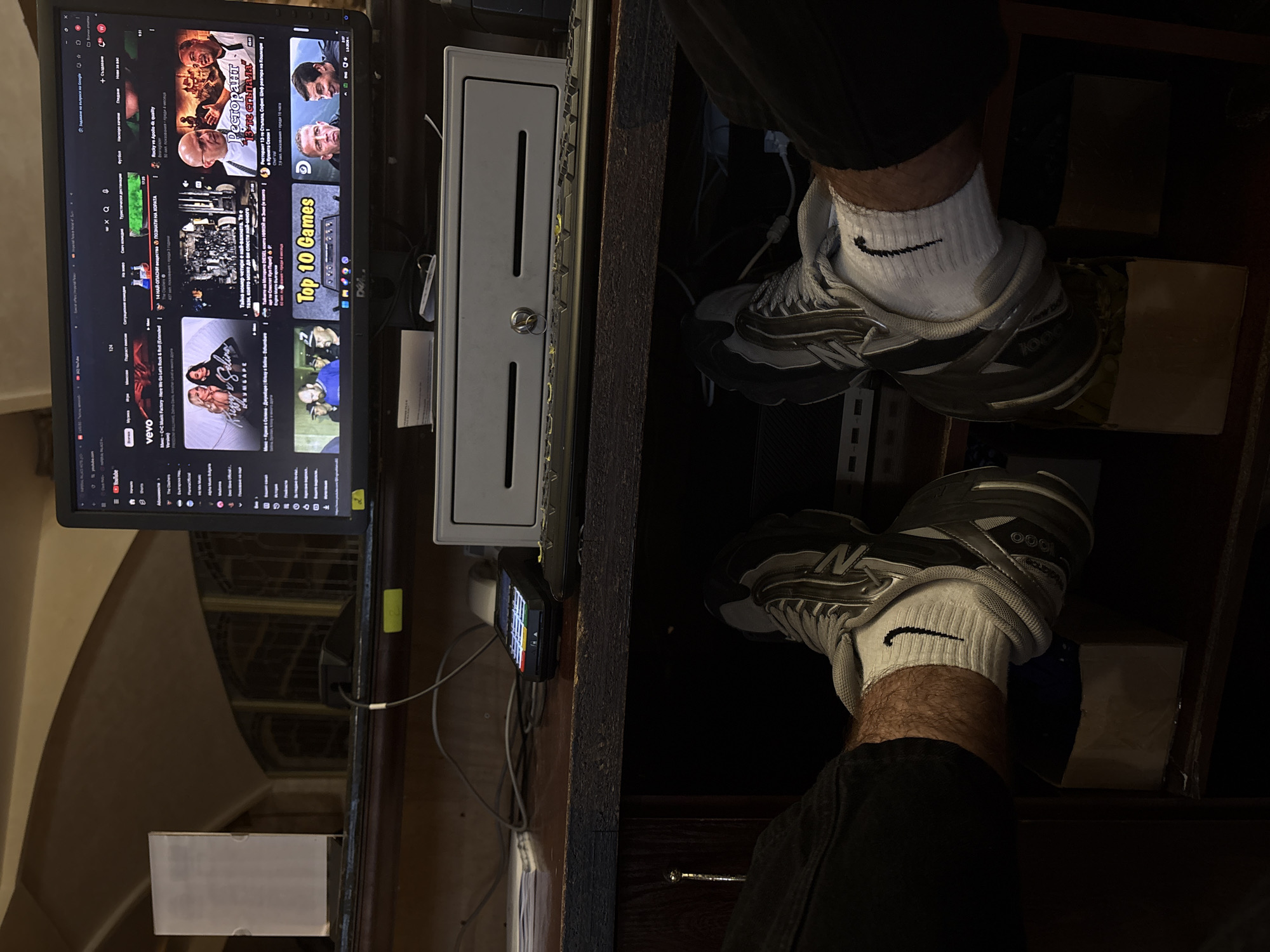 The image shows a person's feet wearing athletic shoes and white socks with a Nike logo, resting on boxes under a desk. The desk is cluttered with electronic devices, including a computer monitor displaying a YouTube interface, a keyboard, and a point-of-sale terminal. In the background, a portion of a building with arched windows and a dark, dimly lit interior is visible. The scene appears to be an indoor workspace, possibly in a commercial establishment. The location is identified as the city of Nesebar, Bulgaria.
