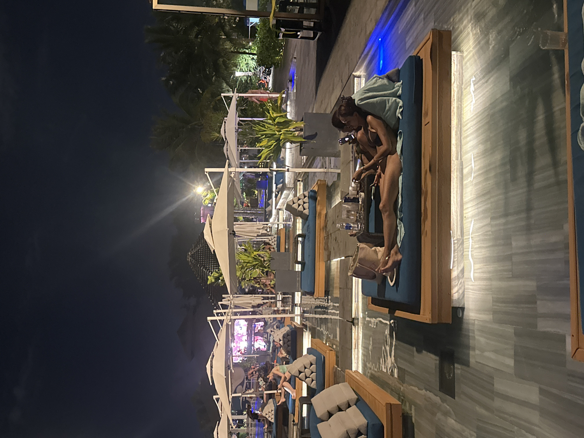 The image depicts an outdoor lounge area at night, likely a bar or resort, in Đà Nẵng, Vietnam. The scene is dimly lit with ambient lighting and some areas illuminated by blue lights, suggesting a relaxed, nighttime atmosphere.
Several individuals are visible, mostly lounging on blue-cushioned daybeds. In the foreground, two women are seated on a daybed. One is looking at her phone, while the other appears to be holding a cigarette. They are dressed in casual attire. Further back, other people are also seen relaxing on daybeds, some appearing to be couples. One person is seen lying down on a daybed in the middle ground. The daybeds are arranged around a pool or water feature, and there are white umbrellas providing shade. Tropical plants are interspersed throughout the area. The ground appears to be tiled with a stone-like pattern that is wet, reflecting the lights. In the background, there are structures with tents or canopies, and glimpses of more populated areas with colorful lights, possibly indicating a resort or entertainment complex.
The time of day is clearly night, and the weather appears to be clear. The presence of palm trees and the mention of Đà Nẵng, Vietnam, suggest a tropical or subtropical location. The overall mood seems to be one of relaxation and leisure. No visible text is present in the image.