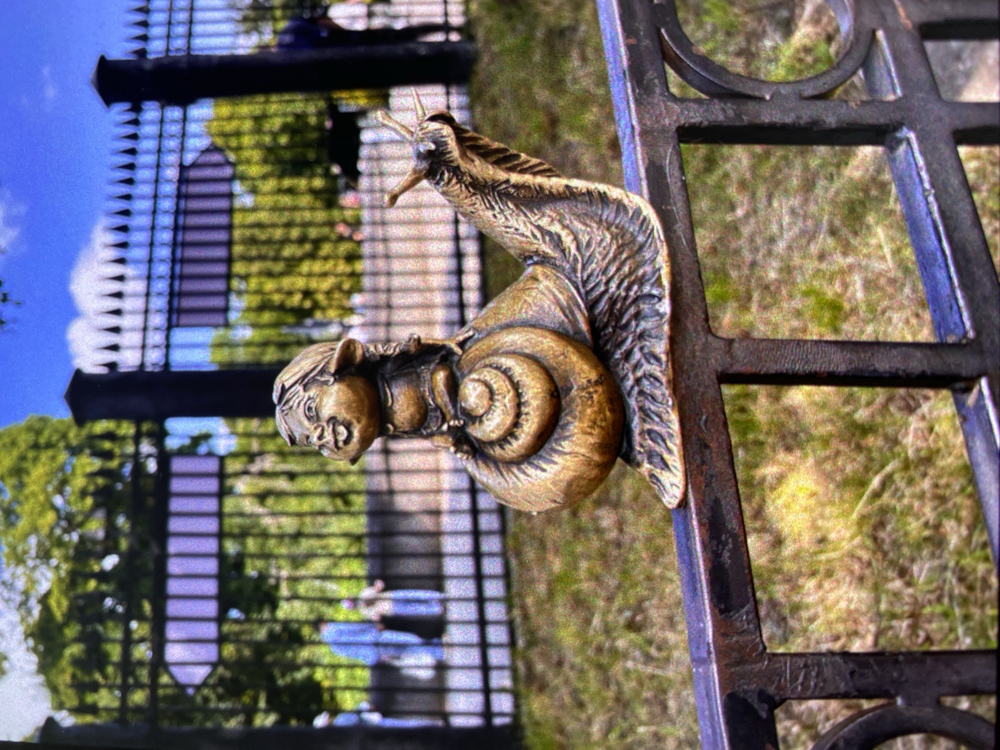 A bronze sculpture of a human-faced snail clings to a black iron fence in Saint Petersburg, Russia. The background features a green lawn and a fenced-in structure.
