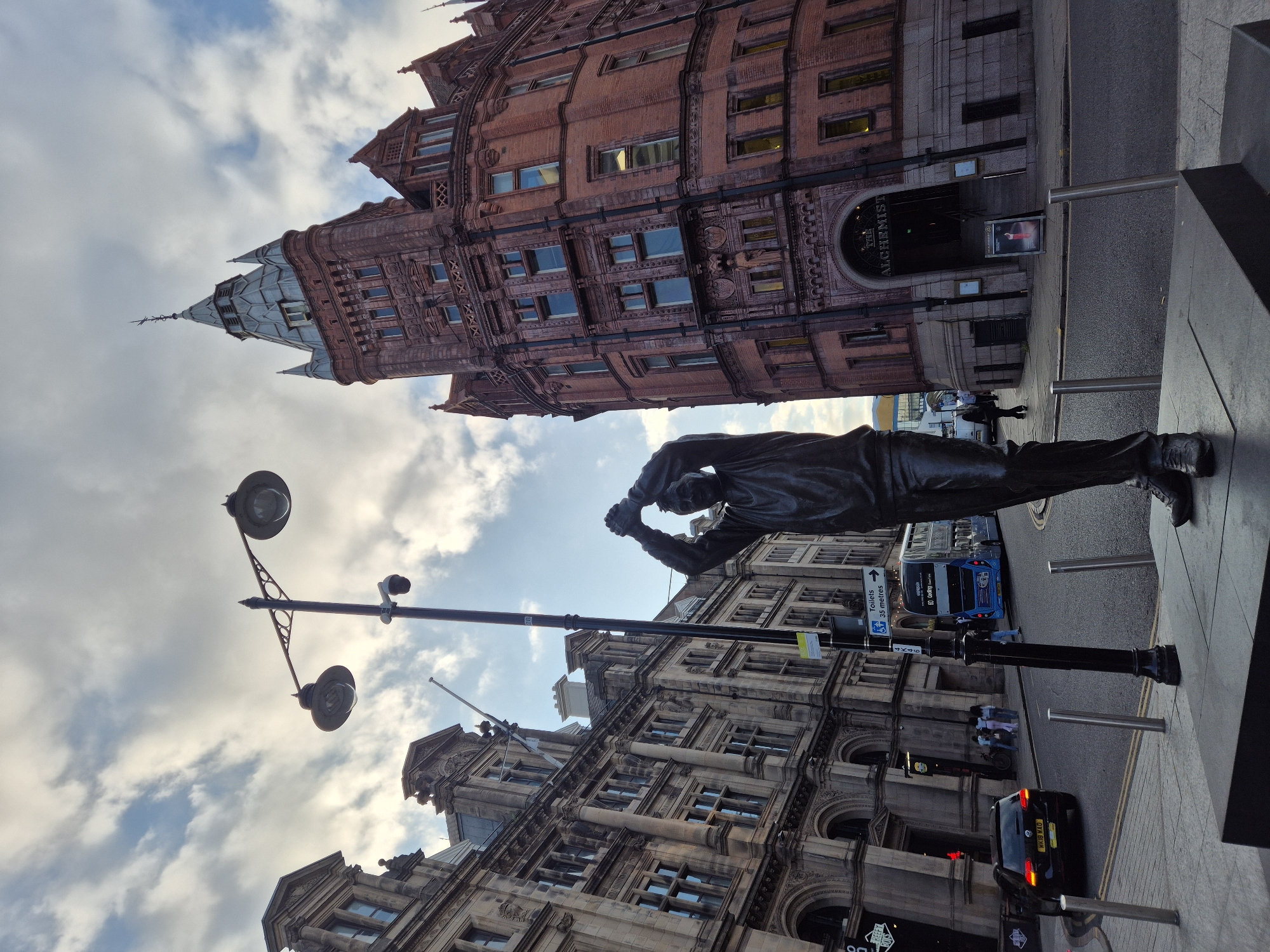 The image shows a street scene in Nottingham, United Kingdom, featuring a statue of a sportsman. The statue, made of dark metal, stands on a grey plinth in the foreground, depicting a man with his arms raised, holding what appears to be a ball above his head. He is wearing athletic attire.
Behind the statue, a street stretches out, with a double-decker bus and a black car visible on the road. To the left, a row of old, ornate stone buildings with many windows lines the street. To the right, a distinctive red brick building with a prominent tower and intricate architectural details dominates the skyline. This building has arched entrances at street level, one of which is labeled "THE ALCHEMIST".
A street lamp with two globes stands near the statue, and a traffic light is visible further down the street. The sky above is partly cloudy, with patches of blue sky visible between the white and grey clouds. Several people are walking on the sidewalk and crossing the street in the background, some near the entrance to the red brick building and others further down the street. The overall impression is a vibrant urban setting with historical architecture.