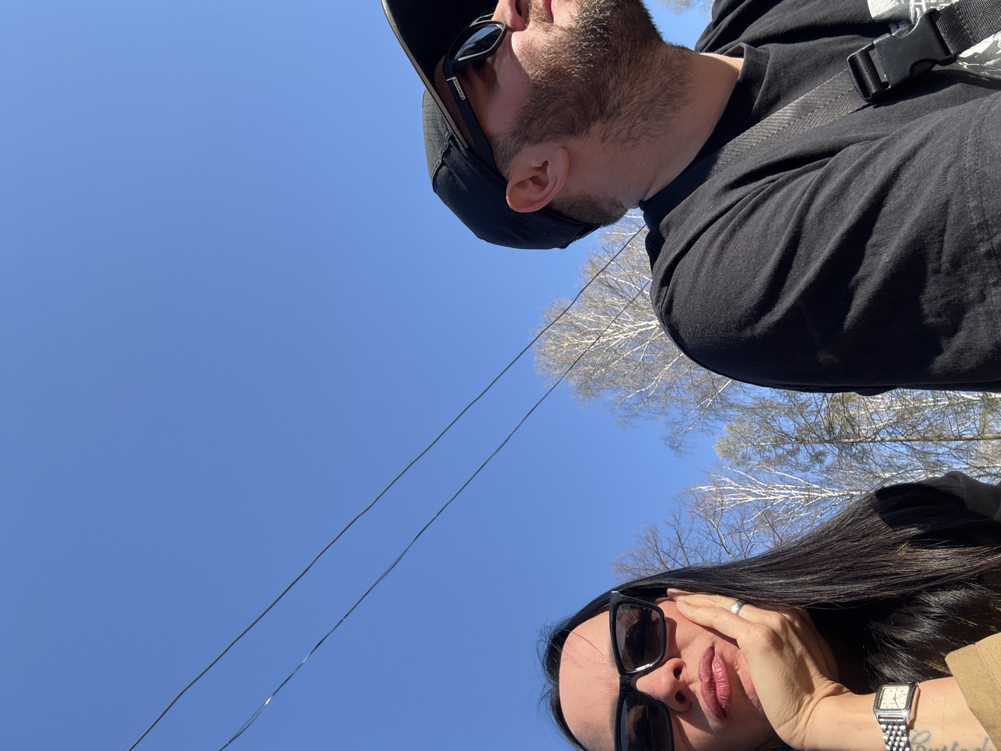 Two people, one man and one woman, are captured from a low angle, looking down at the camera. Above them is a clear blue sky and bare tree branches. The presence of electrical wires suggests an urban setting.
The man, positioned at the top of the frame, has a beard, wears sunglasses, and a black baseball cap. He's dressed in a black t-shirt. A strap is visible across his chest.
The woman, positioned at the bottom of the frame, has long, dark hair. She is also wearing sunglasses. Her hand is near her face. A wristwatch is visible on her wrist.
The location is Chaikovsky, Russia.