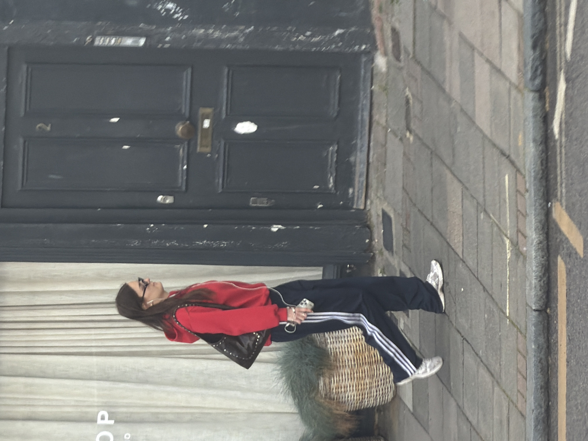 A woman stands on a sidewalk in front of a building. The woman is wearing a red hooded sweatshirt, black pants with white stripes, and white sneakers. She has dark hair and is wearing glasses. She is holding a cell phone in her hand. The building behind her has a dark gray door with the number "2" on it. There is a wicker basket in front of the building. The sidewalk is made of gray bricks. The location is in the city of London, United Kingdom.