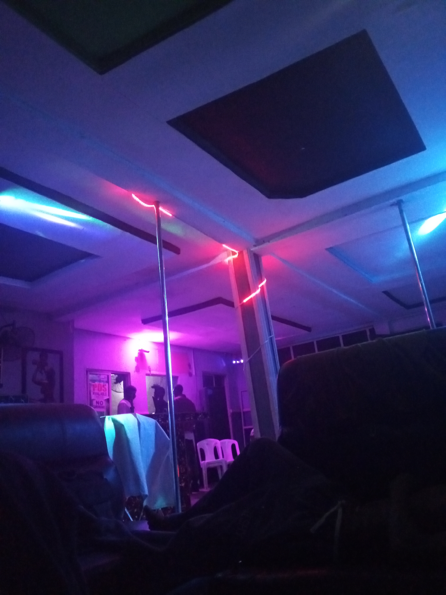 This image depicts the interior of a dimly lit entertainment venue, possibly a club, bar, or lounge. The dominant feature is the ceiling, which is adorned with square recessed panels and illuminated by crisscrossing LED strip lights in vibrant red, blue, and purple colors. These lights cast a colorful glow throughout the space.
Several metallic poles, potentially for performance or structural support, extend from the floor to the ceiling, reflecting the varied hues of the lights. In the foreground, dark upholstered seating, likely a sofa or armchairs, occupies the lower portion of the frame, indicating a seating area for patrons.
Towards the mid-ground, a table draped with a light-colored, possibly white or light blue, tablecloth is visible. Behind it, two white plastic chairs are placed. Silhouettes of several people are gathered near a doorway in the background, with one individual appearing to wear a light-colored shirt. A wall-mounted fan is also visible in this area.
On the wall to the left, framed pictures or posters are discernible but indistinct. Next to the doorway, a sign is partially visible with the text "POS AVAILABLE" and "NO," likely indicating payment options and possibly restrictions.
The scene captures a nighttime setting, given the reliance on artificial and colored lighting. The overall ambiance suggests a lively and social gathering, characteristic of a modern nightlife establishment.