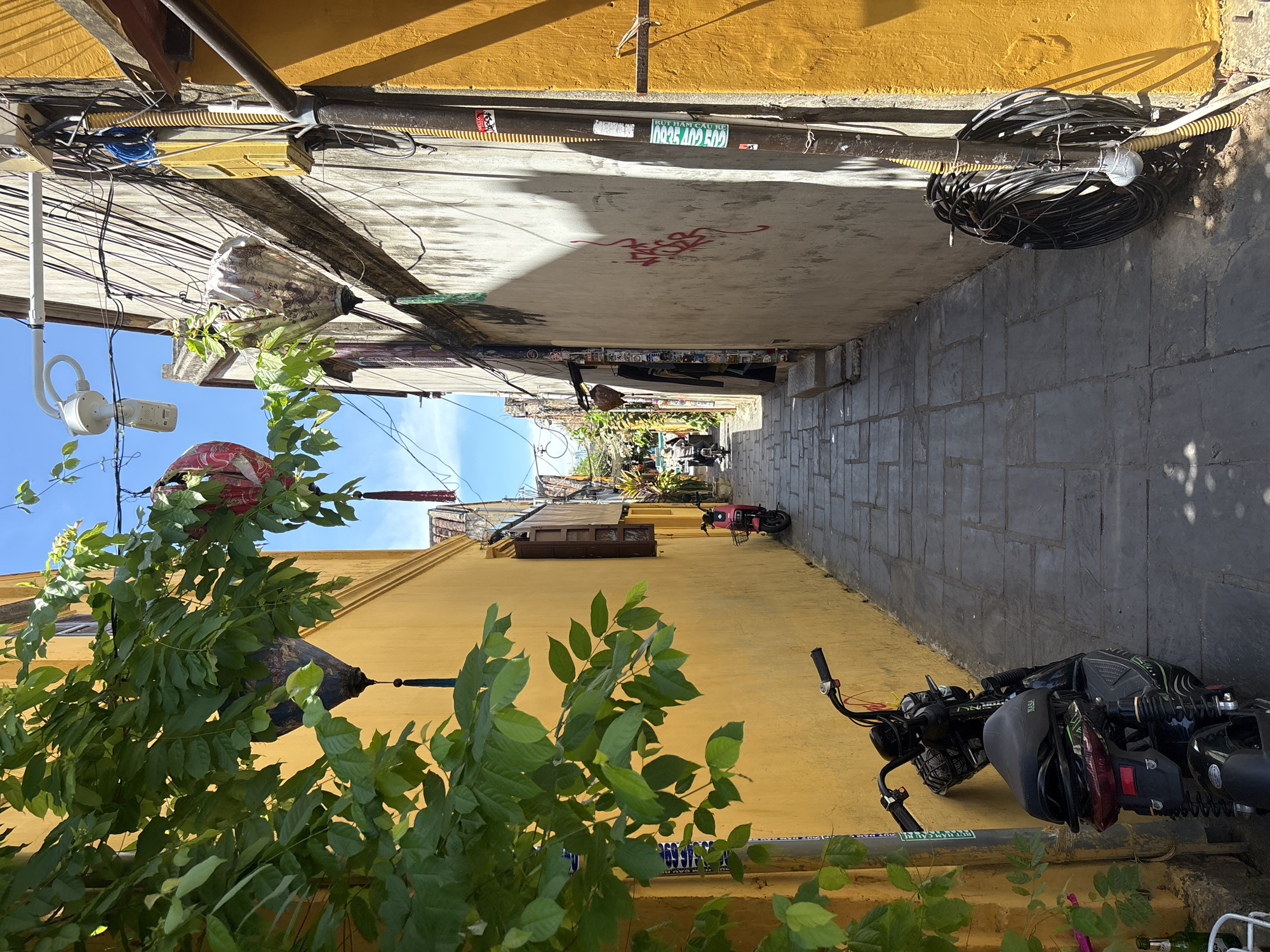 This is a full-frame, eye-level, outdoor, vertical shot of a narrow alleyway in Hoi An, Vietnam. The alley is paved with dark grey flagstones. On the left, a bright yellow building is partially obscured by lush green foliage from a tree with large leaves. Hanging from a branch are two decorative lanterns, one red and white, and another dark metal one with a tassel. A black electric scooter is parked against the yellow wall. On the right, a light-colored building runs along the alley, its wall marked with graffiti and posters. Various electrical wires and conduits are visible running down the side of this building, with a thick bundle of black cables coiled and secured to a pole. A pink electric scooter is parked further down the alley, and a person is seen on a scooter in the distance. The sky above is a clear blue.