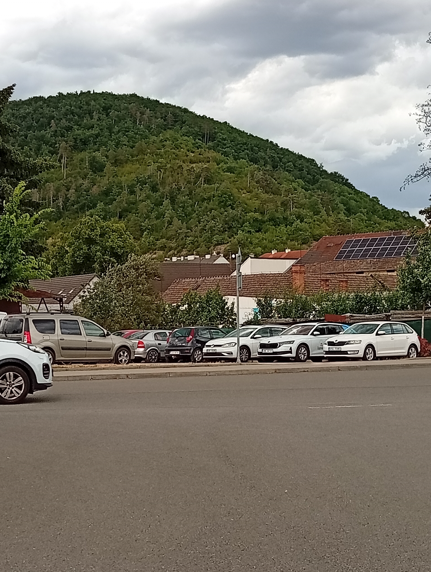 The image shows a street scene in Tišnov, Czechia. A row of parked cars is visible along the side of the road, with a green, tree-covered hill rising in the background. Some buildings with tiled roofs, including one with solar panels, are situated between the road and the hill. The sky is overcast with clouds. The cars are a mix of makes and models, and include a silver SUV, several white hatchbacks, and a black hatchback. There are no people visible in the image.