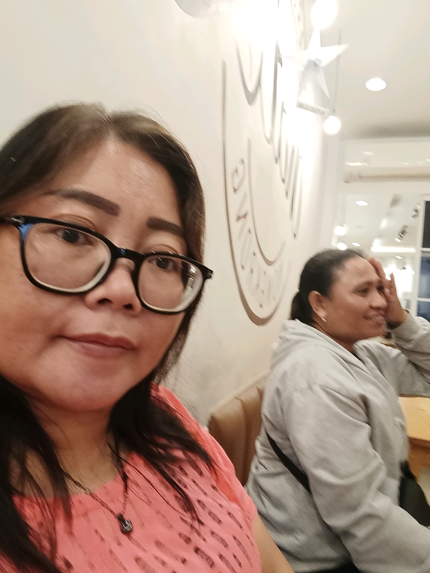 Two adult females are seated inside a well-lit indoor establishment. The female in the foreground, positioned on the left, is closer to the camera and facing it directly. She has dark hair, wears black-rimmed glasses, and is dressed in a pink, textured top with a dark pendant necklace. To the right, a second female is partially visible, facing towards the right side of the frame. She has dark hair pulled back and is wearing a grey hooded jacket. Her right hand is raised to her forehead. The background features a white wall with a large, light-colored circular graphic and multiple ceiling lights. The lighting is bright and even. The overall environment suggests a cafe or restaurant setting in Jatimulya, Indonesia.