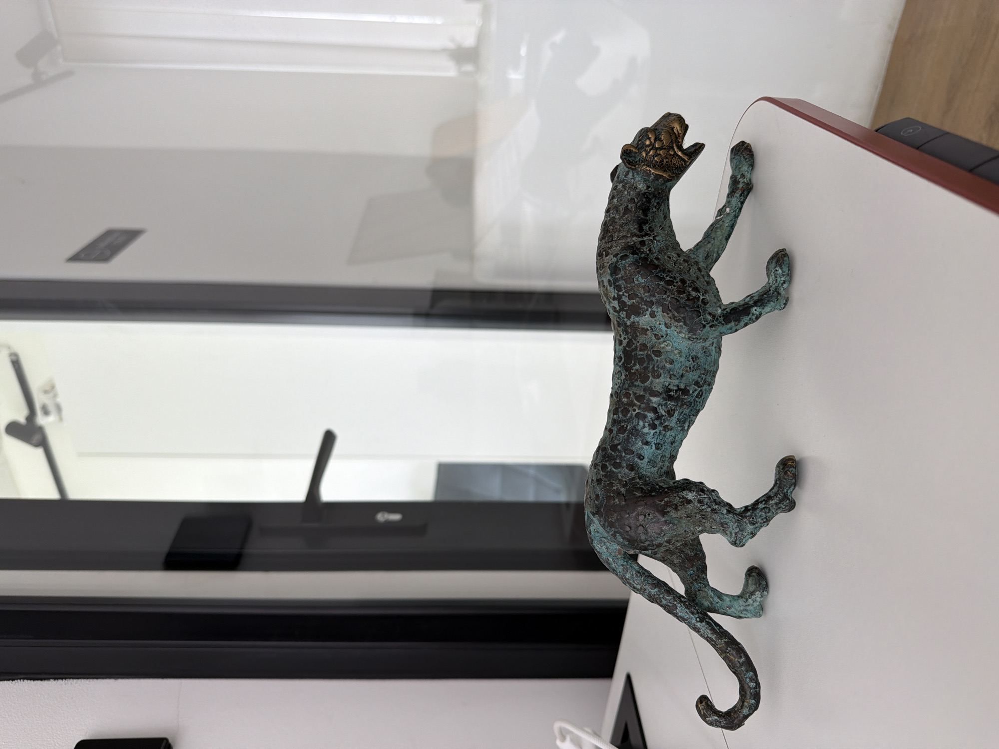 A close-up shot features a bronze or bronze-colored jaguar statue attached to a white surface with a red trim. The jaguar appears to be climbing, with its paws seemingly gripping the surface. The statue exhibits a patina of blue-green oxidation, adding to its aged appearance.
Behind the statue, a window is visible with black framing. Outside the window, a blurred view suggests an exterior environment, though specific details are indiscernible. The image's composition focuses on the statue and its immediate surroundings. The location of North Kuta, Indonesia, is not evident within the visual elements of this image.