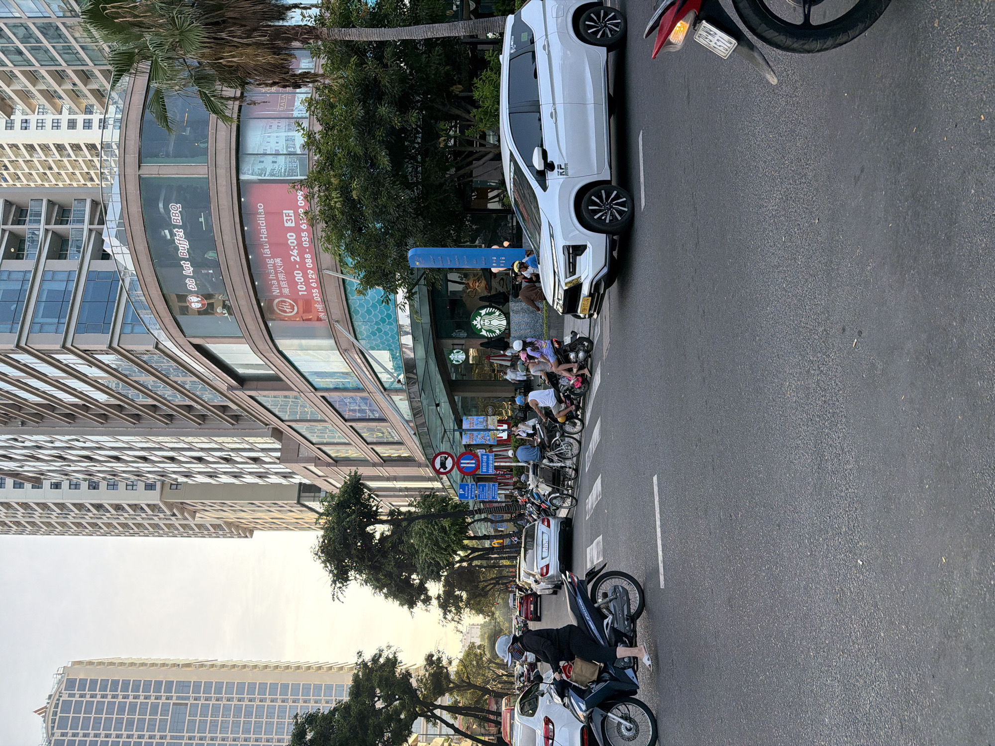This detailed forensic description captures a bustling late-afternoon urban street scene in Nha Trang Ward, Vietnam, characterized by a mix of residential towers, commercial establishments, and heavy traffic. **Location and Environment:** The image depicts a wide asphalt road at an intersection, flanked by multi-story buildings under a clear, bright sky. The warm, soft light with elongated shadows suggests either early morning or late afternoon, likely golden hour. The specific architecture, vehicle types, and signage with Vietnamese text confirm the location as an urban area in Vietnam, specifically Nha Trang based on the prompt and visible "79" prefix on some license plates. Tall, modern high-rise buildings with repetitive window patterns dominate the background on both sides of the street. Palm trees and other broadleaf trees are interspersed along the street and in front of buildings, providing shade and greenery. **Commercial Building:** On the right side of the street, a prominent multi-story commercial building with a curved glass facade is visible. Its ground floor features a Starbucks coffee shop, identifiable by its circular green siren logo and glass entrance. Above Starbucks, a red banner advertises "Nhà hàng lẩu Haidilao" (Haidilao Hotpot), displaying operating hours "10:00 - 24:00" and contact numbers (035 6129 088 - 035 6129 099). Further up, another sign is visible for "Đà Lạt Buffet BBQ." The building's lower levels feature intricate window patterns and a blue, scale-like architectural detail. **Road and Traffic:** The road is busy with a variety of vehicles and pedestrians. White painted lines mark traffic lanes and a crosswalk. * **Vehicles:** Motorcycles and scooters are prevalent, with several parked or in motion. In the foreground on the left, a person wearing a white helmet, face mask, and black attire is seated on a black Honda Future scooter, preparing to proceed. In the mid-ground, a white Mitsubishi Xpander SUV/MPV with license plate "79-AC 113.74" is positioned as if turning right. Behind it, a silver Mercedes-Benz sedan with license plate "79A-431.56" is visible. Multiple other cars, including white and red sedans, are stopped in traffic lanes. On the right side of the road, a traditional Vietnamese bicycle rickshaw (cyclo) with a passenger is visible, alongside other motorcycles. A red motorcycle's rear wheel and license plate "79-N3 1374" are partially visible in the bottom right corner. * **People:** Numerous individuals are present, primarily adults. Many are observed on motorcycles, wearing helmets. Some individuals are walking on the sidewalk near