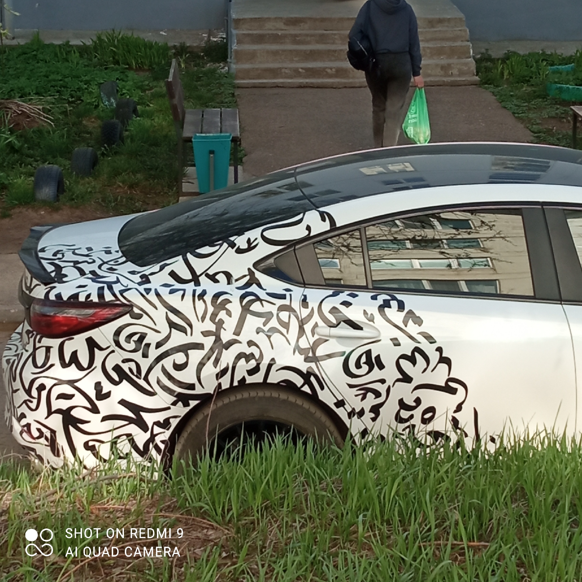 A partial view of a white car with black abstract designs on its side is shown in the foreground. Tall green grass partially obscures the car. Behind the car, in the midground, are a series of steps leading up to a building entrance, where a person walks away from the camera, holding a green plastic bag. A bench and a turquoise waste bin are visible to the left of the steps. In the background, glimpses of the buildings are visible. The image was taken in Chaikovsky, Russia.
