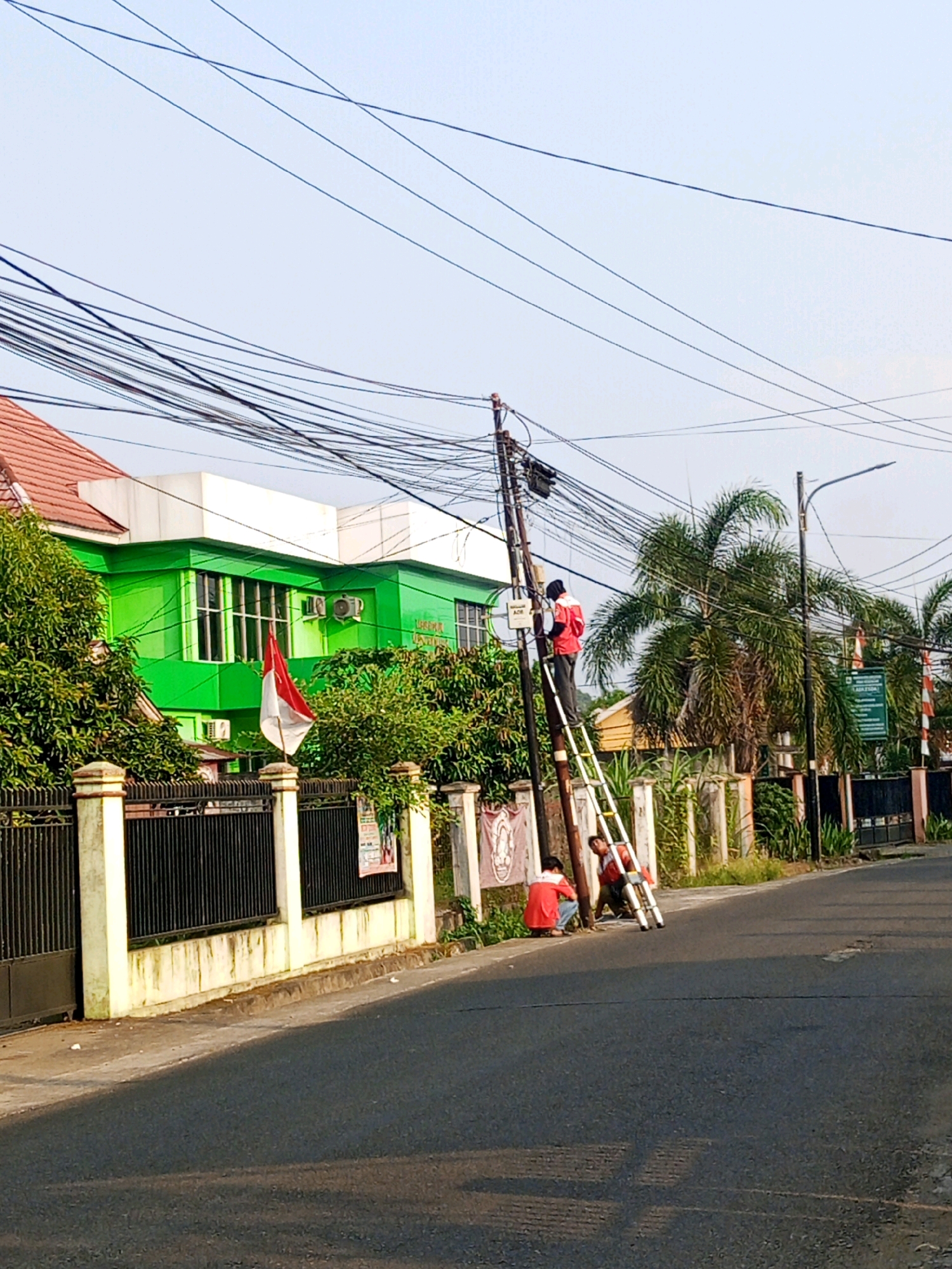 In Lubuklinggau, Indonesia, two individuals are observed working on utility poles along a paved road. One person, wearing a red shirt and black headwear, is positioned on a ladder, actively engaged with the wires. Another individual in a red shirt is seated on the ground next to a second ladder, observing or assisting. The scene includes a green building with a red-tiled roof in the background, a black metal fence, and lush green trees. The sky is clear and blue. A flag, red and white, is displayed on a pole near the building.
