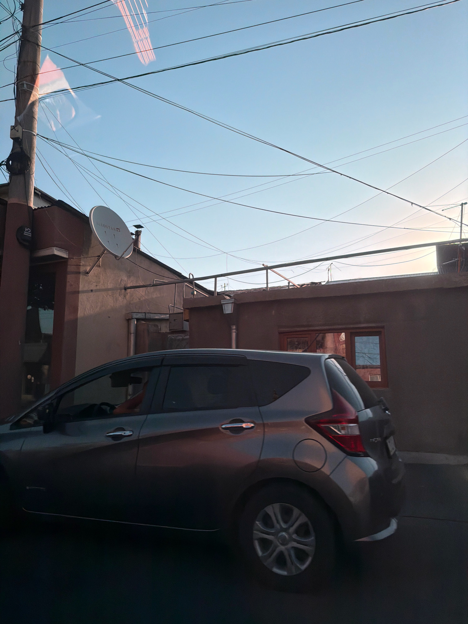 The image shows a grey Nissan Note parked on a street in Yerevan, Armenia, against a backdrop of buildings and a clear blue sky. Overhead, a tangle of electrical wires crisscrosses the sky, supported by a wooden utility pole on the left. A satellite dish is mounted on the side of one of the buildings. The car is viewed from the side, with its rear wheel and tail light visible. The setting sun casts a warm glow, reflecting off the car's surface and the building's walls, creating subtle highlights and shadows. The overall scene conveys a sense of everyday urban life in Yerevan.