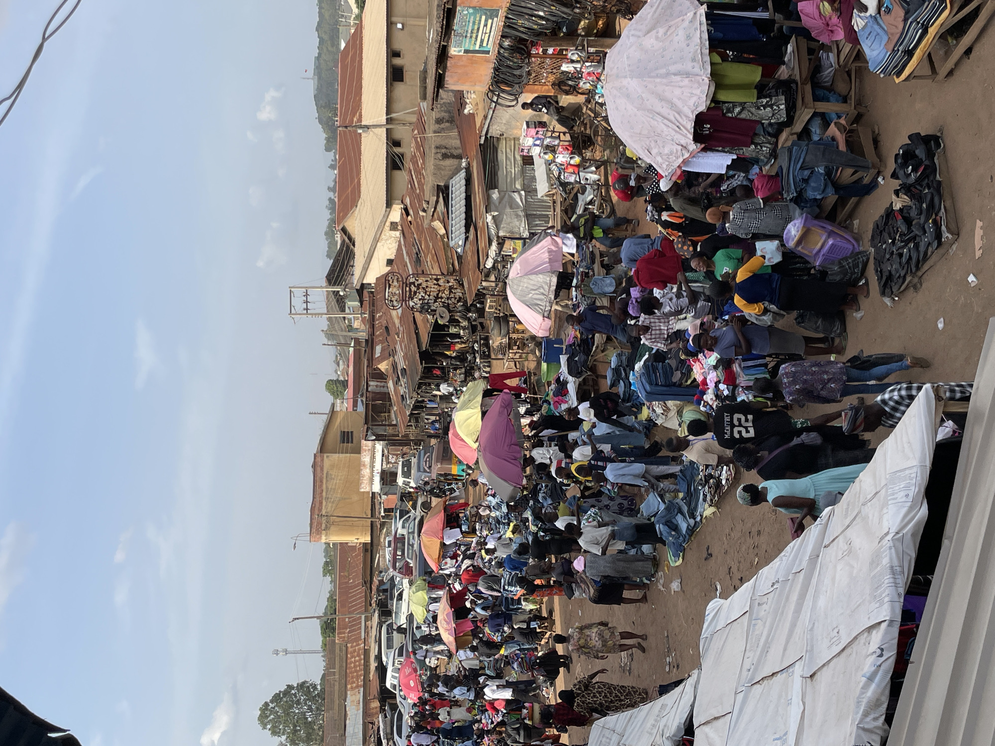 The image depicts a crowded outdoor market in an unidentified city, with a high density of people and goods. Vendors are displaying items, possibly clothing and other merchandise, on the ground and on elevated surfaces like tables. Several individuals are using umbrellas for shade. The buildings in the background have a weathered appearance with corrugated metal roofing. The ground is a mix of earth and possibly paved areas. The overall environment suggests a bustling commercial area.