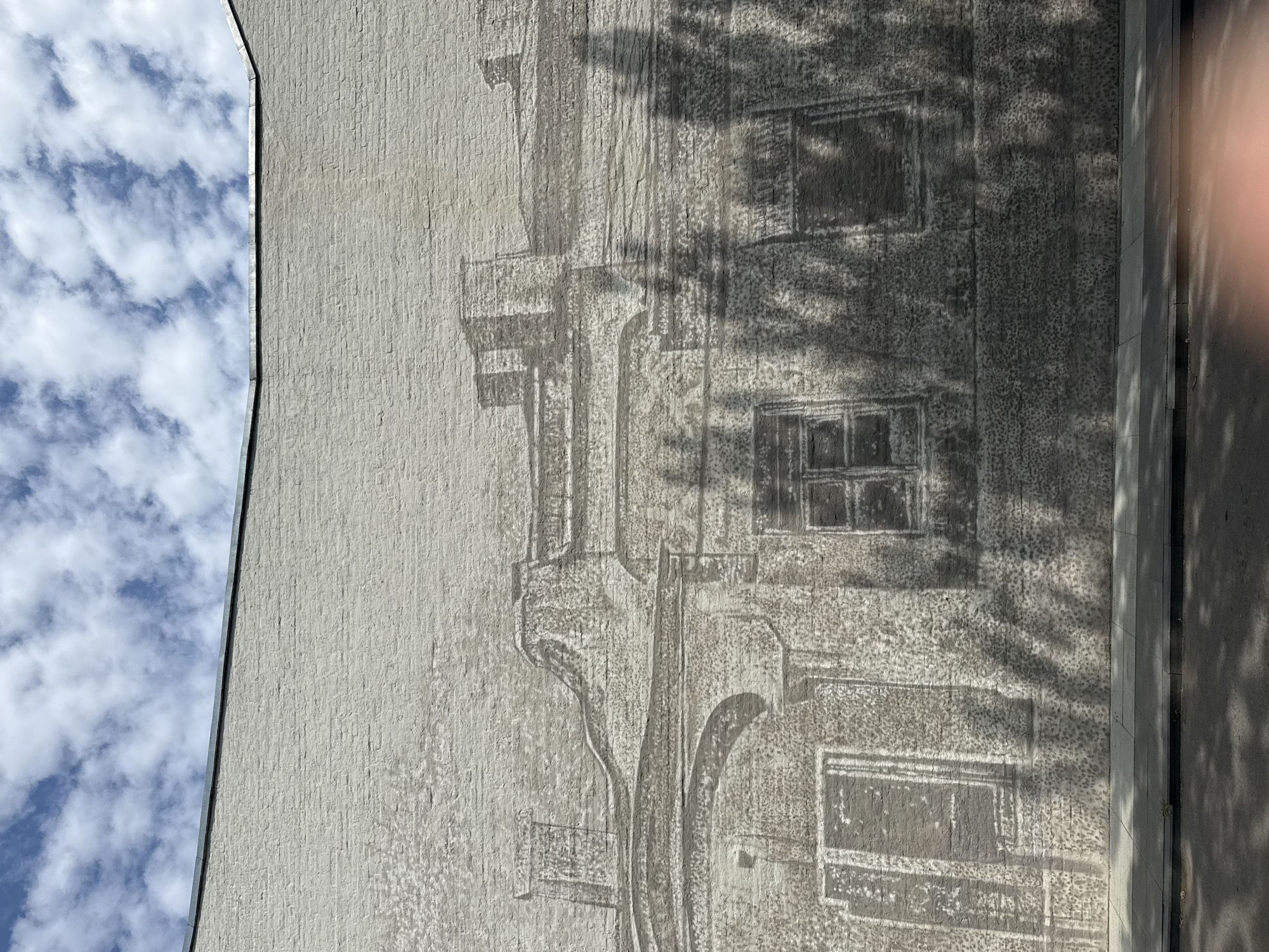The image shows a mural on a brick wall, depicting a sketch of a building with chimneys and windows. The mural is rendered in shades of gray and blends with the texture of the bricks. The top of the wall meets a clear blue sky filled with white, fluffy clouds. A metal coping runs along the top edge of the wall. In the foreground, there is a sidewalk and a paved road, with shadows of trees cast across both. There are no people or animals visible in the image. The scene appears to be an outdoor urban environment. While the image does not explicitly state the location, the architectural style of the mural could be found in many historic city centers.
