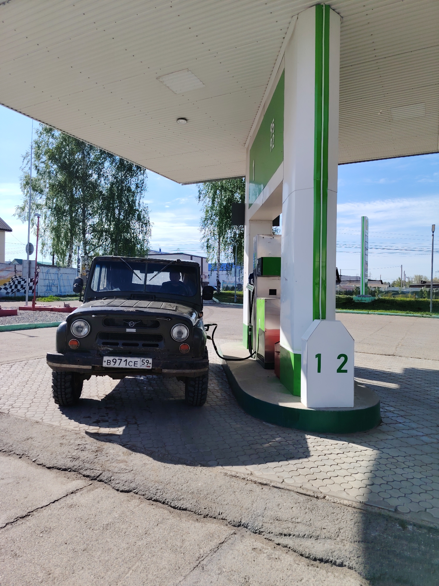 The image depicts a UAZ-469 jeep parked at a gas station in Chaikovsky, Russia. The vehicle, painted a dark color, is positioned in front of a gas pump with the number "12" prominently displayed. The license plate of the UAZ-469 reads "B971CE 59 RUS". The gas station has a white and green color scheme. Trees and other buildings are visible in the background under a partly cloudy sky. A person is visible inside the UAZ-469.