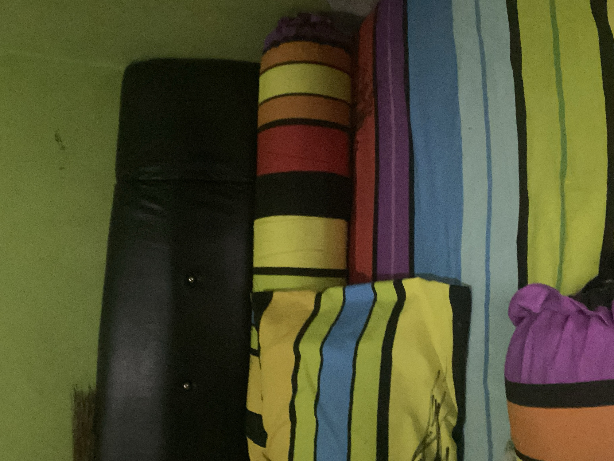 A close-up, eye-level shot shows a bed with a black headboard and a brightly colored, striped comforter. The comforter has horizontal stripes of yellow, green, blue, purple, and orange, with black lines separating the colors. A cylindrical bolster pillow, also striped, lies on the bed. The wall behind the bed is painted a light green color. The scene appears to be indoors, likely a bedroom, and no specific location in Ta', Indonesia, can be determined from the image alone.