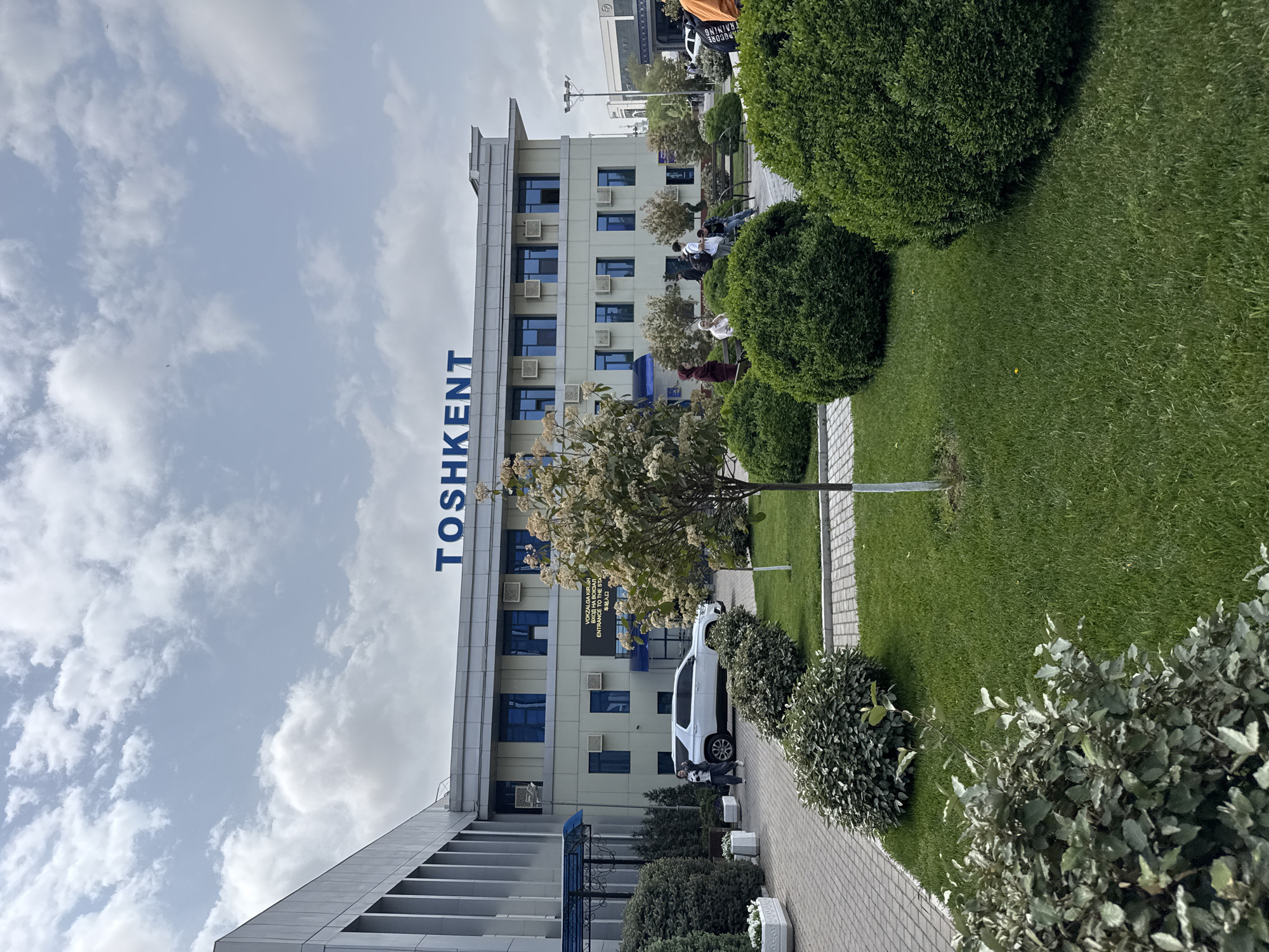The photograph depicts a building with the word "TOSHKENT" written vertically in large blue letters. The building has a modern architectural style with multiple floors and blue-framed windows. Air conditioning units are visible under some windows. A white car is parked next to the building.
There is a paved pathway leading along the side of the building, bordered by manicured green lawns and various bushes and small trees. The sky is partly cloudy.
The location is Tashkent, Uzbekistan, as indicated by the building signage. People are present in the photo.