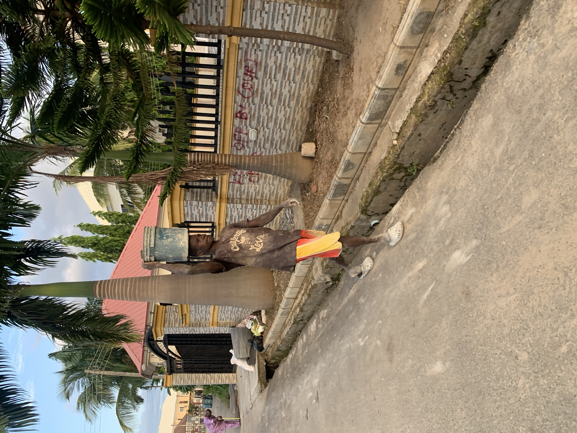 A man in a brown t-shirt, red and yellow shorts, and white sandals is carrying a bucket on his head while walking on a street in Abuja, Nigeria. The street is paved and has a curb and a ditch. Palm trees and buildings surround the street. Two people in purple outfits are standing in the distance. A building wall with black fencing and spray-painted writing is visible.