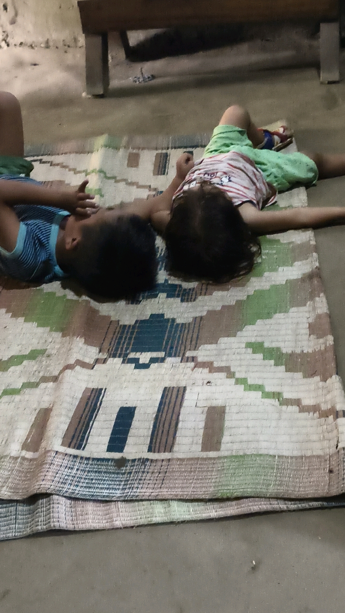 Two children are lying on a woven mat on a concrete floor. The child on the left is wearing a blue and white striped shirt and appears to be a boy. The child on the right is wearing a white shirt with red and blue stripes and green shorts. This scene is located in the city of Mualsang, India. A dark wooden bench is visible in the background, casting a shadow on the floor. The mat has a geometric pattern of brown, green, and blue stripes.