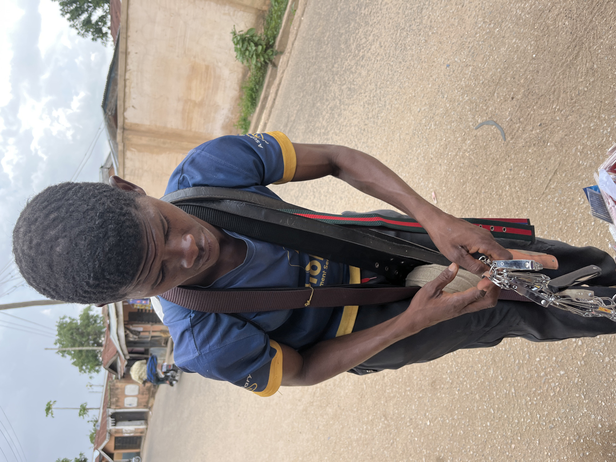 A man stands on a paved road in Jos, Nigeria, holding a collection of metal tools and implements. He is facing downwards, examining the tools in his hands. The man is wearing a dark blue t-shirt with yellow accents on the sleeves and a dark-colored strap across his torso. Behind him is a low beige wall with a patch of vegetation at the top. Other buildings can be seen in the background, with trees interspersed. The sky is overcast.