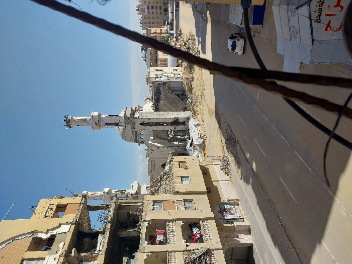 The image displays a scene of significant urban destruction in Khan Yunis, Palestinian Territory, under a clear blue sky. In the midground, a prominent white mosque with a tall, ornate minaret and a central dome is visible, exhibiting substantial structural damage, including collapsed sections and surrounding piles of concrete rubble. To the left, a multi-story, light-beige building stands heavily damaged, with several floors partially collapsed, exposing internal structures and twisted rebar. Laundry, comprising red and white items, hangs from a balcony on this damaged building. A paved two-lane street traverses the scene, extending from the foreground into the midground, with large accumulations of debris and rubble flanking its edges. A single white hatchback car is parked on the right side of the street, facing away from the viewer. In the far background on the right, additional multi-story residential buildings appear relatively intact. Dark, indistinct structural elements partially obstruct the immediate foreground, suggesting the photograph was taken from an elevated position within another structure. No individuals are discernible in the scene.