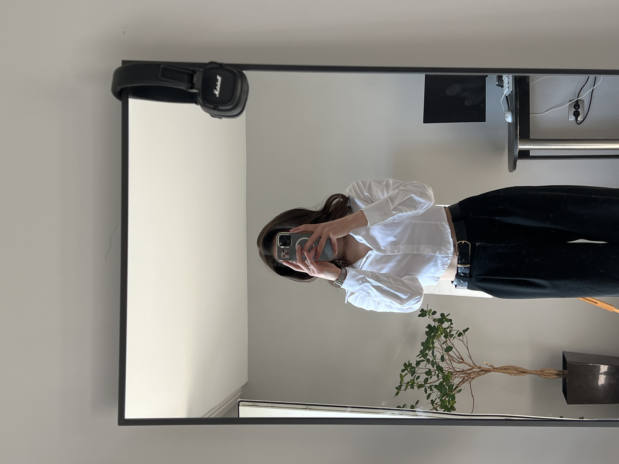 A person stands in front of a large mirror, taking a selfie. They are wearing a white, cropped button-down shirt and black high-waisted pants with a black belt. Their hair is dark and falls to their shoulders. In the reflection, a potted plant is visible to the left, and a small desk with a computer monitor, keyboard, and mouse is to the right. A pair of black headphones is hanging from the top of the mirror frame. The room has light grey walls. The image was taken in Ternopil, Ukraine.