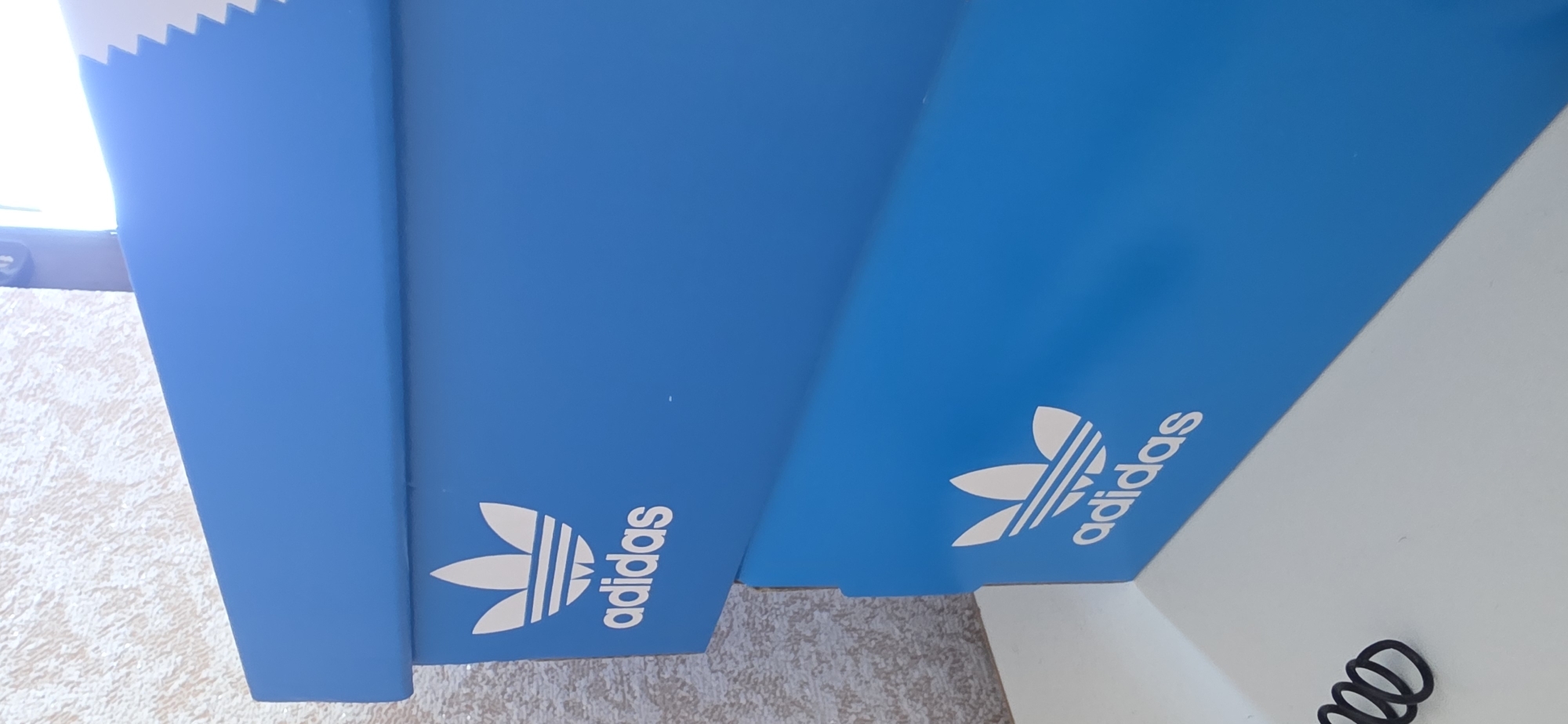 Two stacked blue Adidas shoeboxes are shown. The top box has the Adidas logo and brand name printed in white on its side. The bottom box also has the Adidas logo printed in white. The boxes are placed on a white surface, which appears to be a shelf or table. In the background, a textured wall is visible, with a lighting fixture at the top right. To the left of the bottom box, a coiled black object, possibly a cable or string, is present on the white surface. The scene is indoors. The image does not contain information about the city of Sumgayit, Azerbaijan.