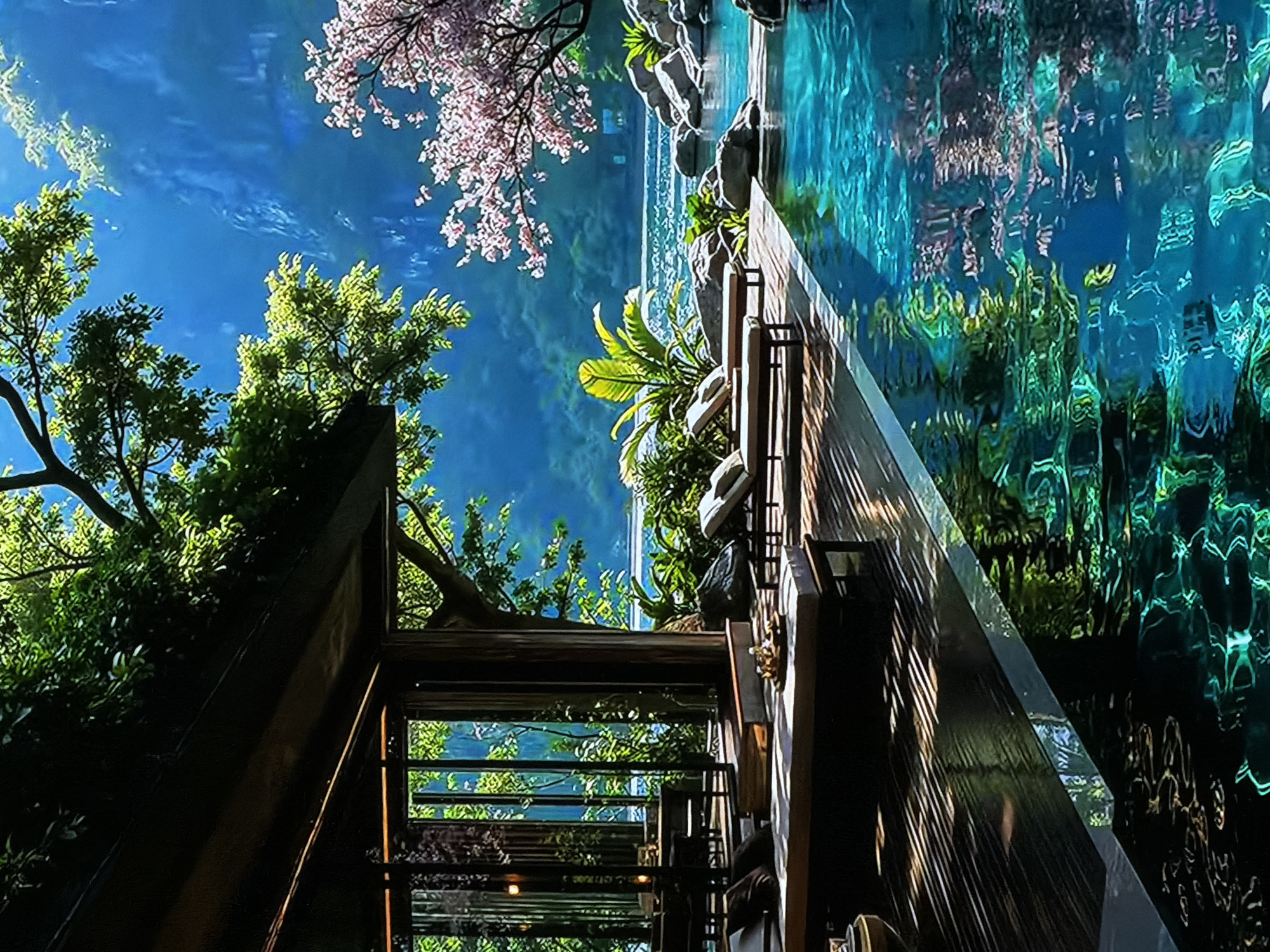 This vivid and artfully composed image, likely depicting a scene in Sofiivska Borshchahivka, Ukraine, captures a serene, lush environment characterized by clear waters, abundant greenery, and thoughtful architectural elements. The perspective looks upward and across a vibrant landscape.
A prominent feature is a large body of luminous blue-green water on the right, shimmering with reflections of the sky and surrounding foliage, creating intricate, abstract patterns. The water's edge is adorned with natural rocks and tropical-looking green plants, suggesting a well-maintained aquatic feature.
On the left, a dense canopy of various green trees, with leaves brightly illuminated by sunlight, fills the frame. In the upper part of the scene, delicate pink blossoms, resembling cherry or almond flowers, cascade from a branch against the clear blue sky, adding a touch of spring beauty. The sky itself is a bright, unblemished blue with subtle wisps of white.
Dark-toned architectural structures, possibly wooden or metal, cut across the middle ground. These appear to be parts of a walkway, balcony, or building, featuring railings and structural beams. Integrated transparent panels, likely glass, reflect the vibrant surroundings, further enhancing the interplay of light and color. One large, diagonal glass panel creates a striking reflective surface.
The scene conveys a peaceful, almost dream-like atmosphere, suggesting a meticulously designed garden, an upscale resort area, or a grand botanical display. The bright, clear lighting indicates daytime, likely a sunny morning or afternoon. No human figures or discernible text are present, allowing the focus to remain on the harmonious blend of nature and modern design. The overall impression is one of tranquility and natural splendor.