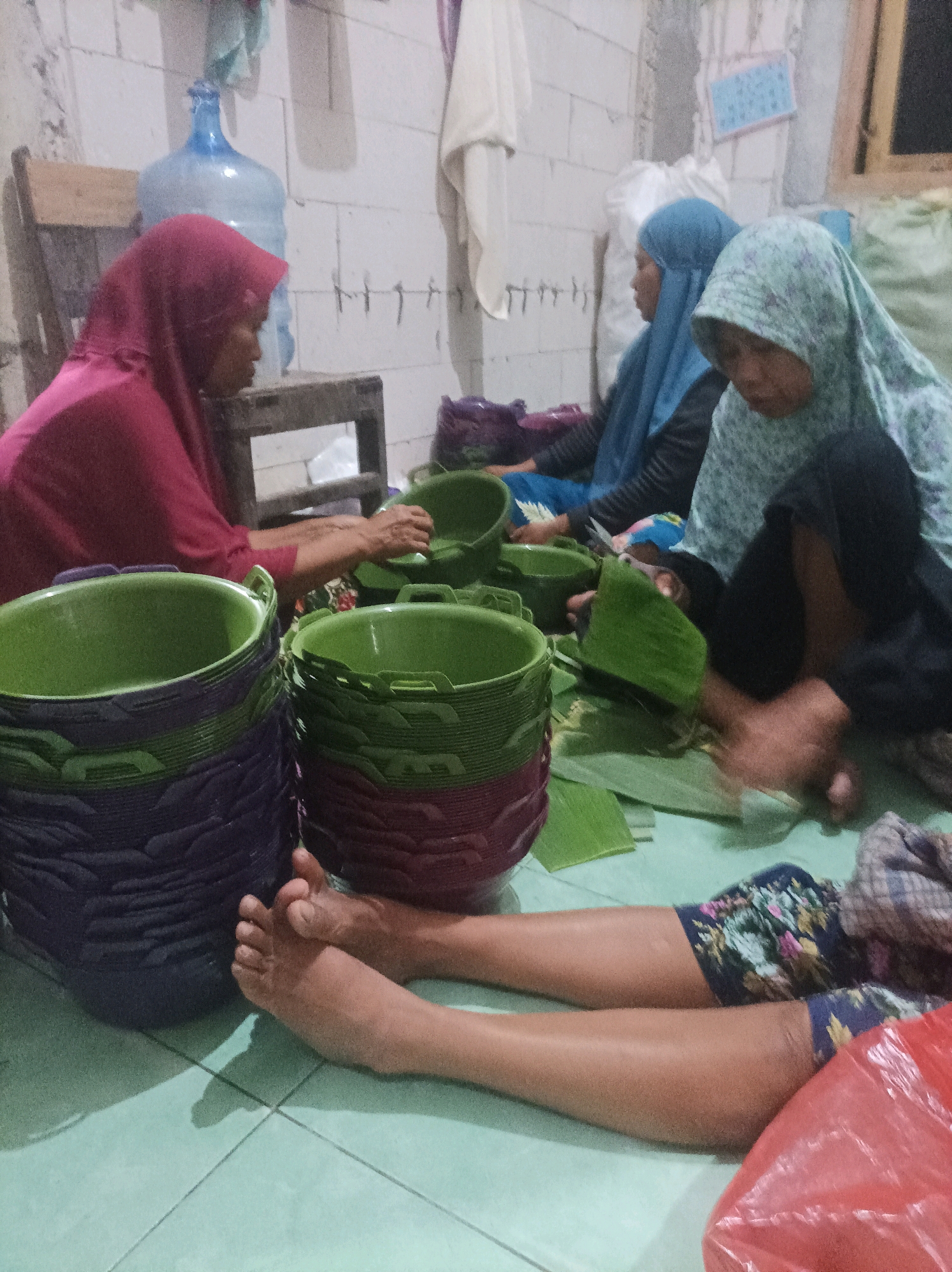 In CANGA'AN, Indonesia, three women are engaged in preparing food in what appears to be a residential setting. Two women are wearing hijabs; one in pink and the other in blue. The third woman is partially visible and wearing a light-colored hijab. They are surrounded by stacks of large green and purple plastic buckets. Banana leaves are spread out on the floor, and the women are seen cutting and arranging them. One woman is holding scissors, indicating they are preparing the leaves for some purpose, possibly for wrapping food. The floor is tiled in a light turquoise color, and the walls are made of white cinder blocks. A large blue water dispenser is visible in the background. The scene suggests a communal activity related to food preparation, possibly for a gathering or event.