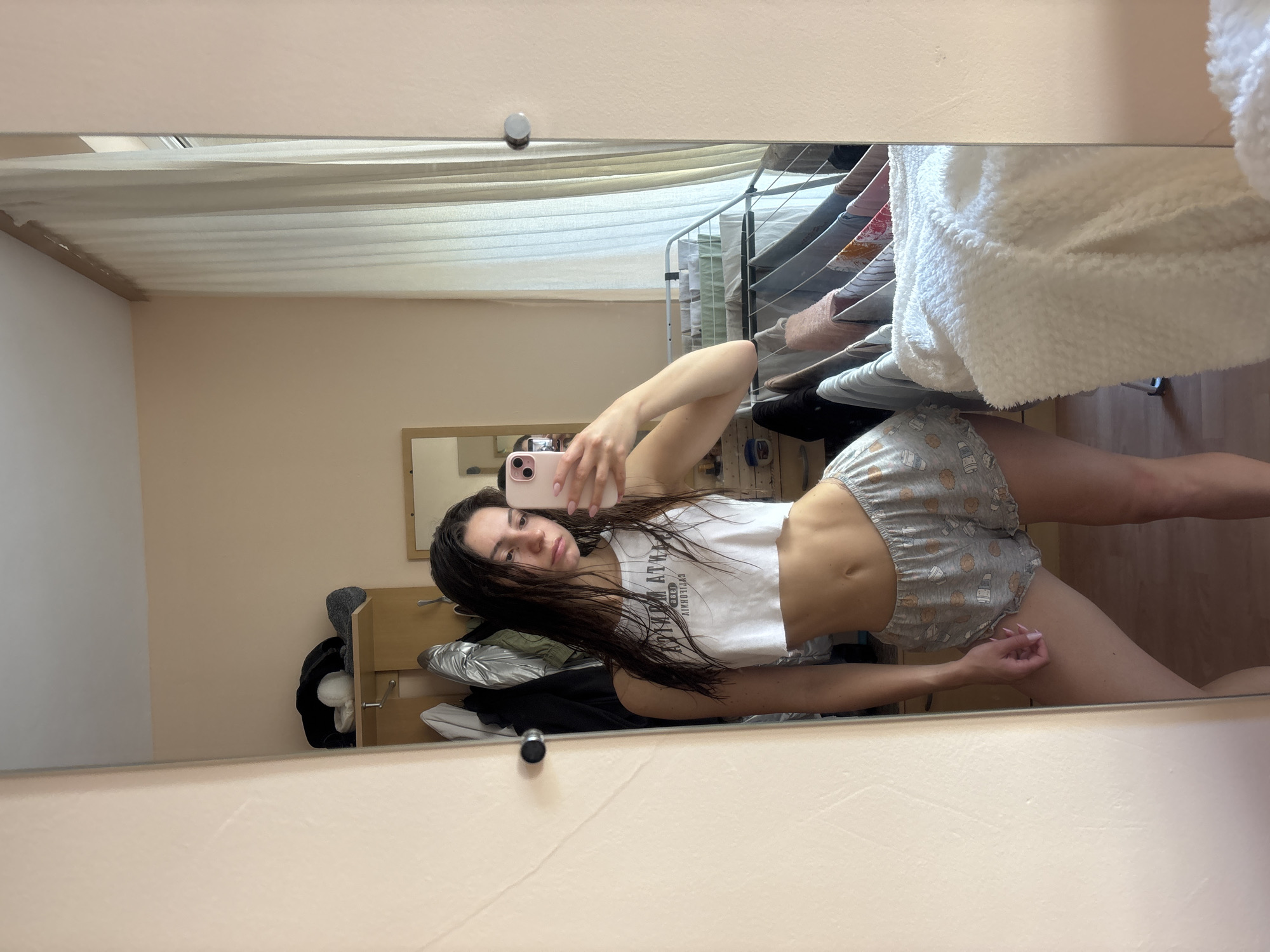 A young woman takes a mirror selfie in what appears to be a domestic setting in Varna, Bulgaria. She is reflected in a large, full-length mirror, showcasing a casual pose where she leans back, extending one leg. She holds a pink-cased smartphone with her right hand to capture the image.
The woman has long, dark hair, and wears a white sleeveless crop top with visible reversed text that reads "URBAN ATHLETICS" and possibly more below it. She pairs this with light grey shorts patterned with various small, cartoon-like designs, including what look like bears and bottles. Her midriff is visible, and she has light pink nail polish.
In the background, visible through the mirror's reflection, are light-colored (beige/peach) walls and a ceiling. To the right, a white clothes drying rack with various items of clothing, including a white fluffy blanket, is present. A smaller, wooden-framed mirror is also visible on a distant wall. On the left side of the frame, reflected behind the woman, is some wooden furniture, possibly a shelf or small cabinet, with clothes or other items piled on it. The lighting is soft and even, suggesting daytime. The overall scene conveys a relaxed, casual moment at home.