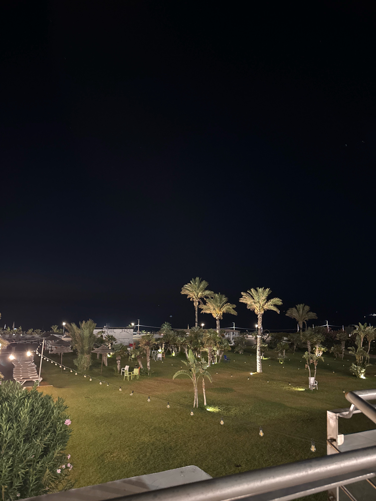 The image shows an outdoor area at night, likely a resort or a public space, with a clear, dark sky. The foreground features a railing and a lush green bush with pink flowers. Beyond this, a well-maintained lawn is illuminated by numerous small lights, creating a festive ambiance. Several palm trees of varying heights are scattered across the lawn, some with their fronds lit from below. In the mid-ground, there are tables and chairs, some with umbrellas, suggesting an outdoor seating area. A structure labeled "POOL BAR" is visible in the distance. In the far background, the silhouette of palm trees and structures can be seen against the dark horizon, with faint lights indicating a built-up area or a harbor. The overall scene is serene and suggests a place for relaxation and entertainment in the evening. The specific location in Isola delle Femmine, Italy, is not explicitly identifiable from the image content alone, but the presence of palm trees and a coastal setting could be consistent with many Italian coastal towns.