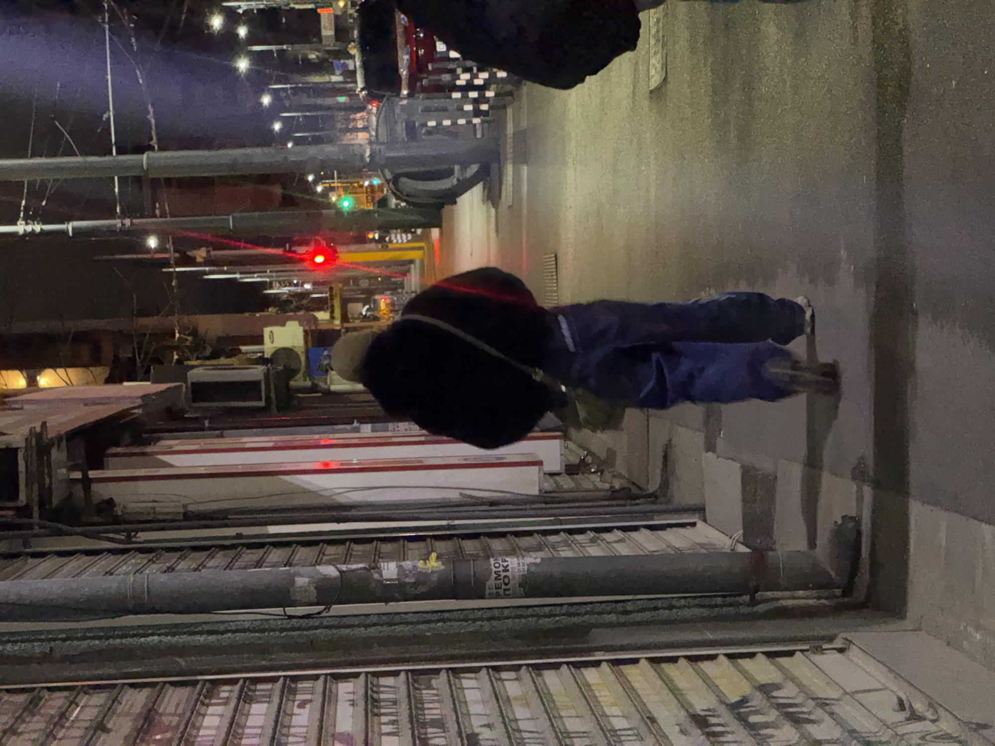 The image depicts a street scene in Sofia, Bulgaria, at night. A person wearing a dark jacket and blue jeans is walking on the sidewalk. In the background, there are shops with closed metal shutters. Overhead are streetlights and traffic lights, one showing green and another showing red. The street appears wet, likely due to rain. A metallic pipe runs along the side of the buildings.