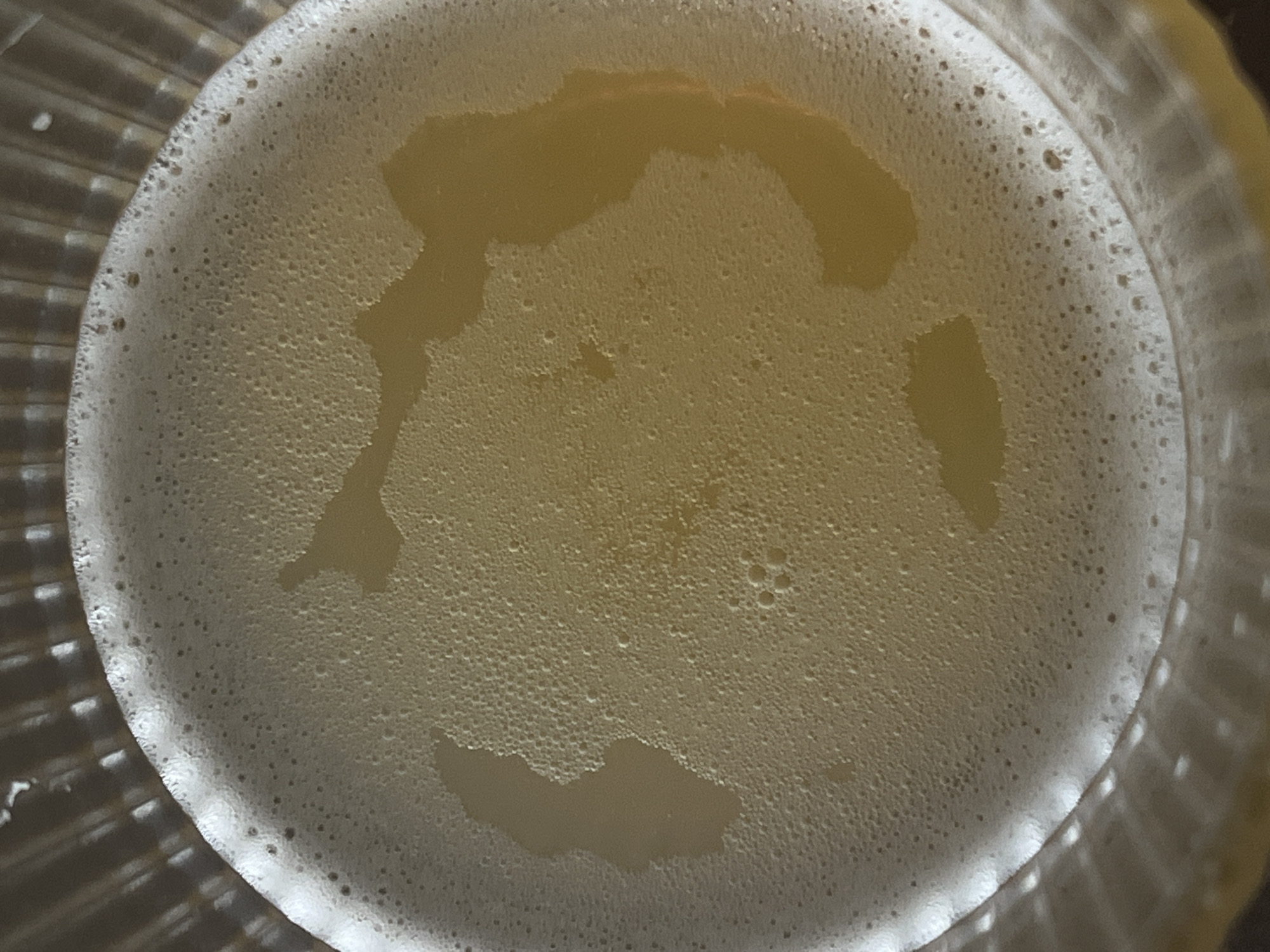 The image shows a close-up, top-down view of the foamy head on a beverage, likely beer, in a clear, ribbed glass. The foam is light yellow and uneven, with some larger bubbles and areas where the liquid underneath is visible, creating darker patches. The glass's textured rim is visible at the bottom and edges of the frame. The scene is illuminated by overhead light, casting soft shadows and highlighting the texture of the foam. The image is an abstract view of a drink and does not depict any specific location in Genemuiden, Netherlands.