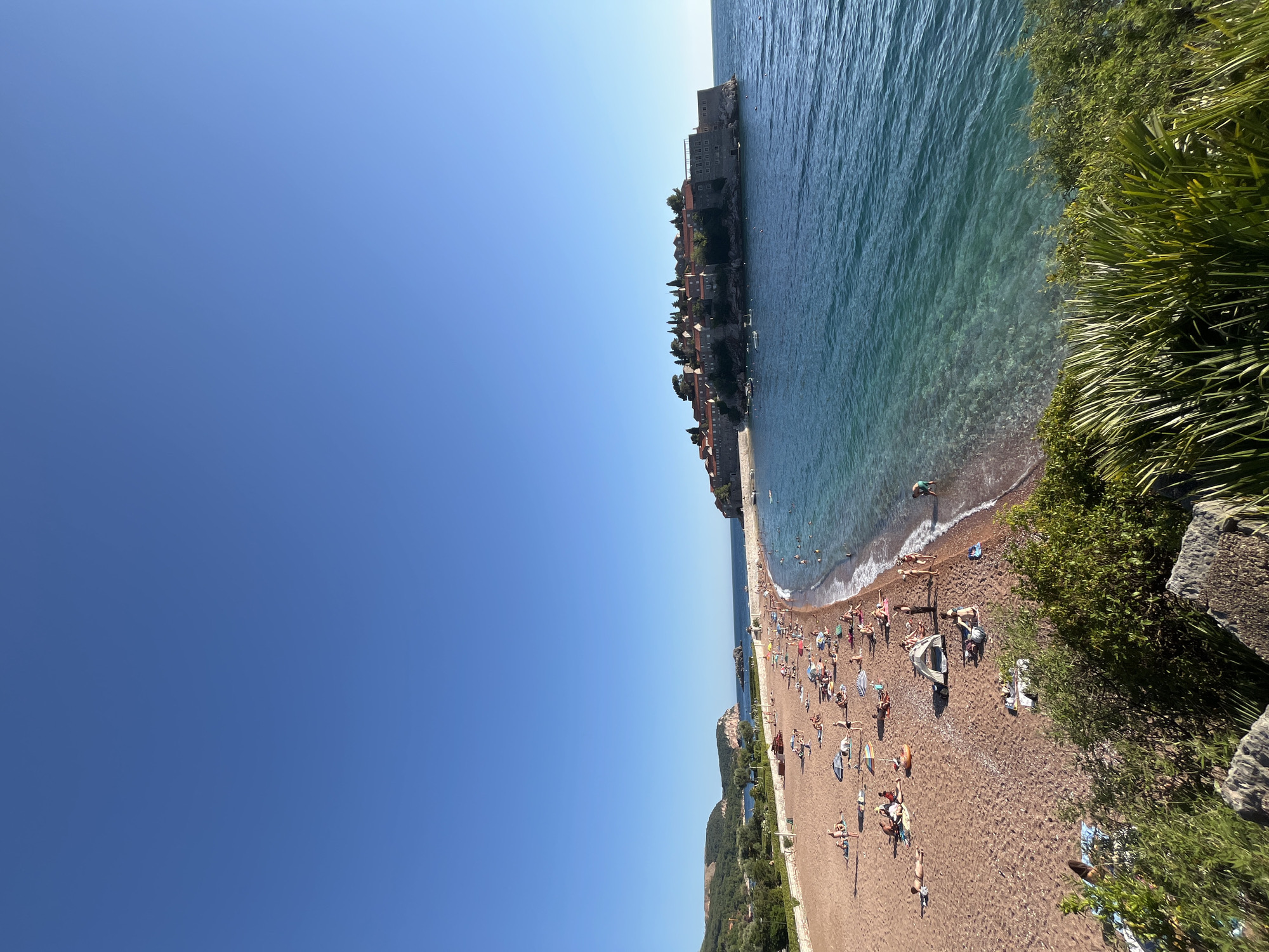 This image depicts a panoramic view of the coastline near Boreti, Montenegro, featuring the island of Sveti Stefan and its adjacent beach. The sky is clear and a uniform bright blue, occupying the upper half of the frame.
In the mid-ground, the prominent feature is Sveti Stefan island, a rocky outcrop on the right side, densely covered with traditional red-tiled roof buildings and green trees. The island is surrounded by deep blue-green water, and several small boats are visible anchored around its perimeter.
A crescent-shaped, reddish-brown pebble beach stretches from the left mid-ground towards the island. The water immediately adjacent to the beach is turquoise and transparent, revealing the pebbly seabed. Gentle waves are breaking along the shoreline. Numerous people, approximately fifty to sixty, are present on the beach and in the water. They are engaged in typical beach activities, including sunbathing on towels and lounge chairs, relaxing under striped and plain beach umbrellas, and swimming or wading in the sea. A small, light-colored tent is pitched on the sand towards the center-right of the beach.
Behind the beach on the left, green, tree-covered hills rise, with some distant structures visible nestled within the foliage. A low, light-colored stone wall runs parallel to the beach, separating it from a vegetated area to the left. The immediate foreground, at the bottom of the image, consists of dark rocks and dense green foliage, including large, palm-like leaves, indicating an elevated perspective.